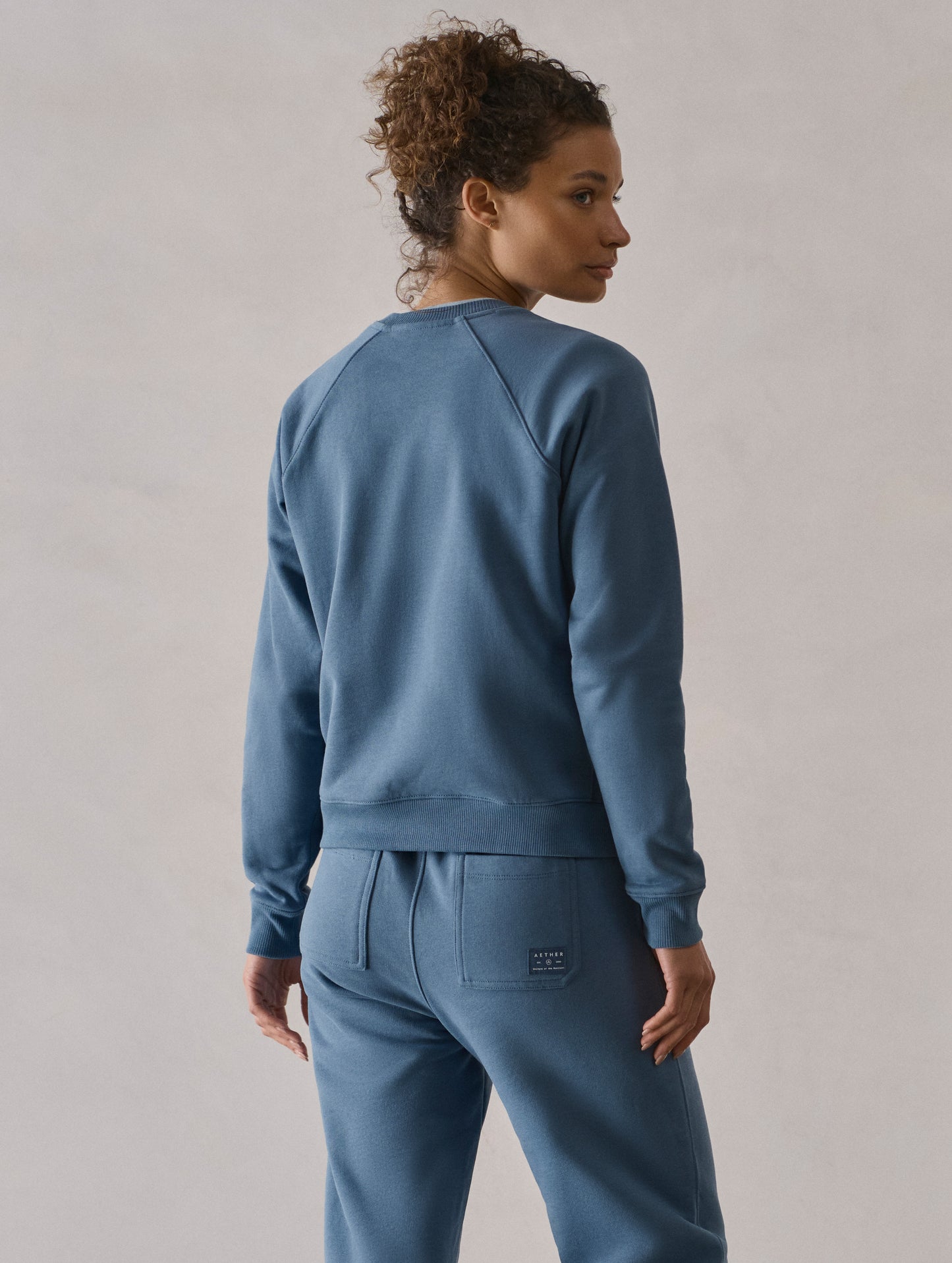 Rear 3/4 upper-body view of woman wearing W Osa Raglan Sweatshirt in Coastal Blue from AETHER Apparel.