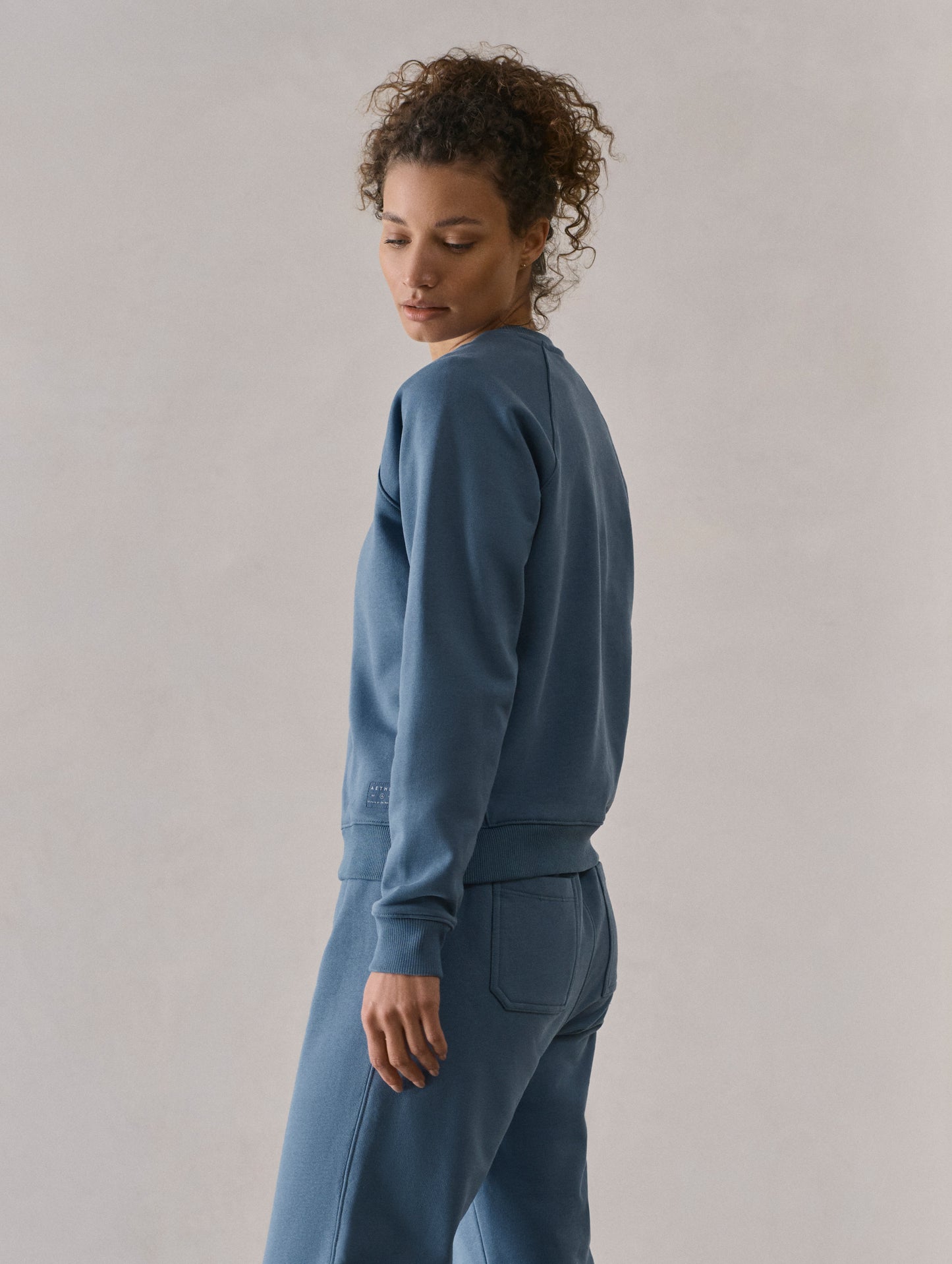 Left side profile upper-body view of woman wearing W Osa Raglan Sweatshirt in Coastal Blue from AETHER Apparel.