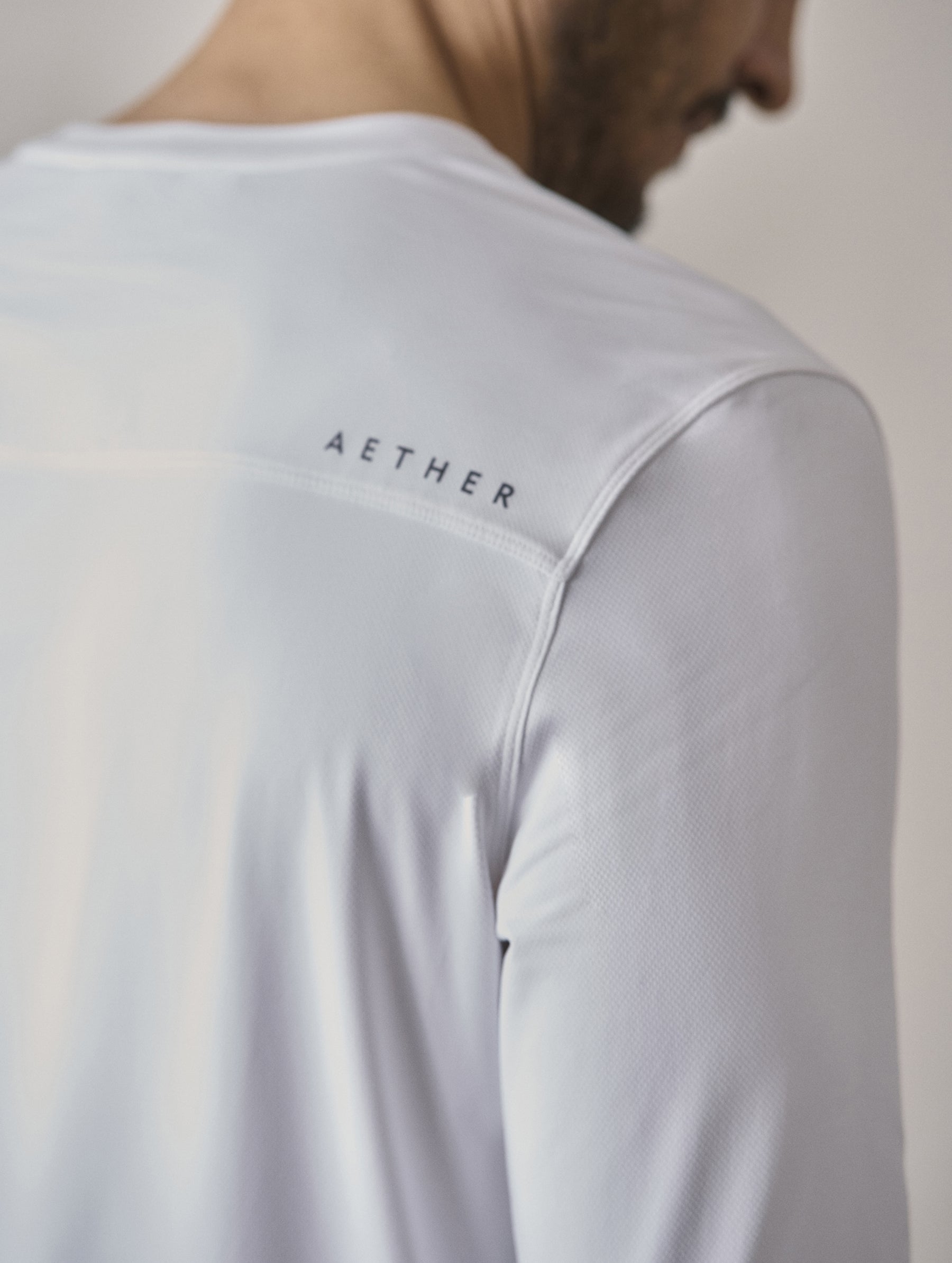 Closeup detailed view of silicone AETHER wordmark on wearer’s back right on the Adventure Performance Long-Sleeve Tee in White from AETHER Apparel.