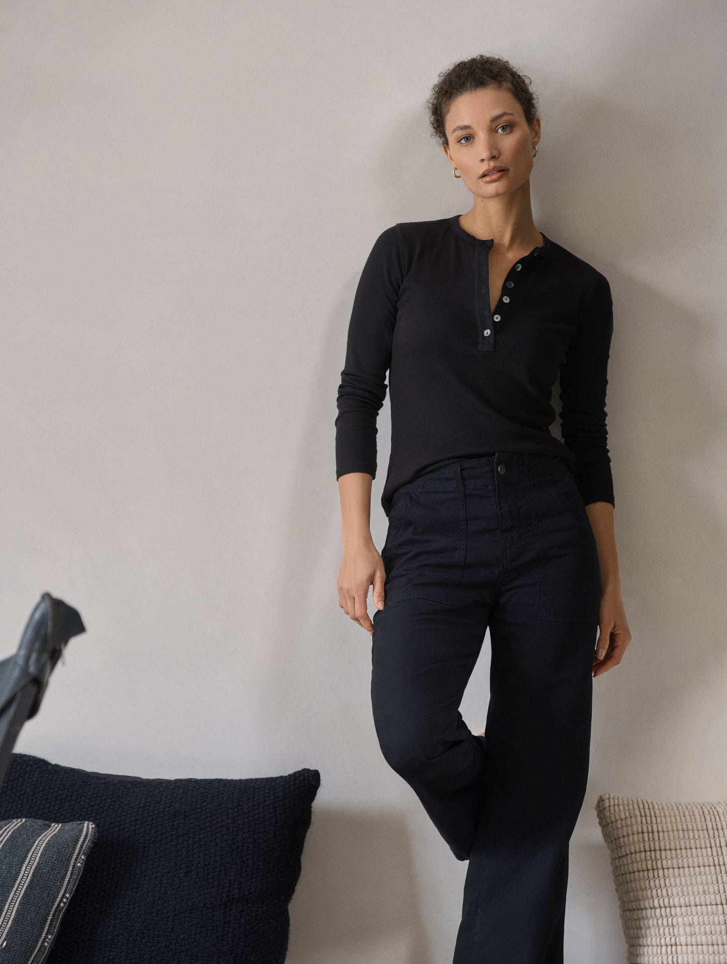 Lifestyle image of woman leaning against wall wearing Amara Long-Sleeve Ribbed Henley in Onyx Black from AETHER Apparel.