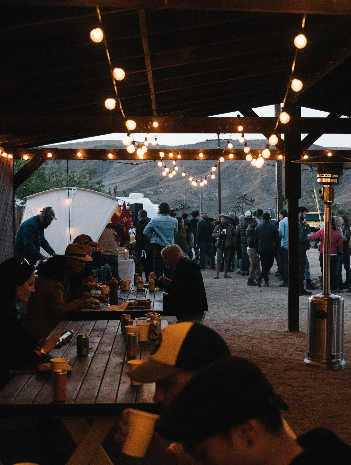 Image of Pioneertown Rally attendees eating dinner on benches under a roof and hanging lights at the AETHER Pioneertown Rally.