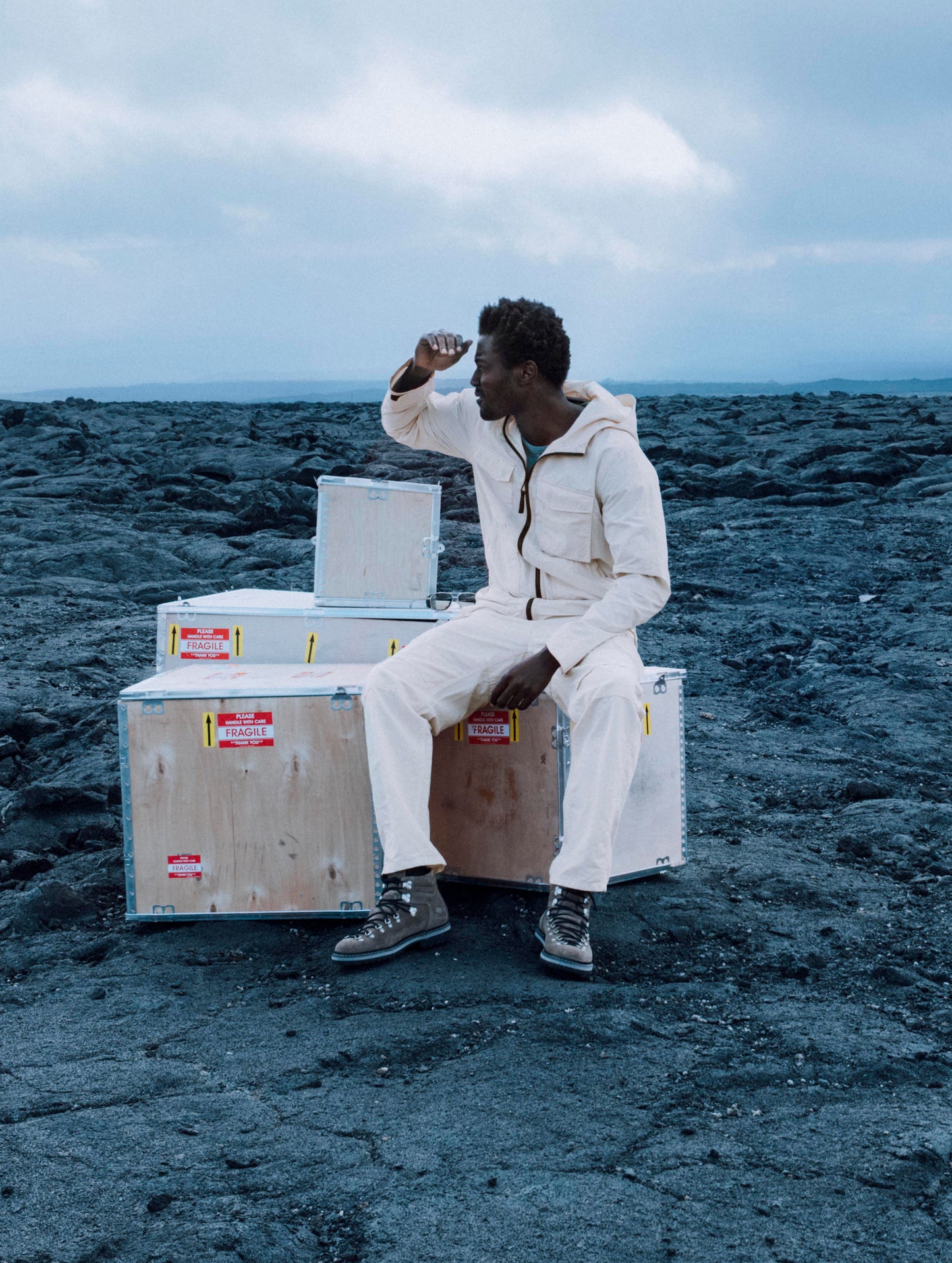 Lifestyle image of a man sitting on top of wooden crates in a lava field while wearing the Aires Lightweight Windbreaker in Bone white from AETHER Apparel.