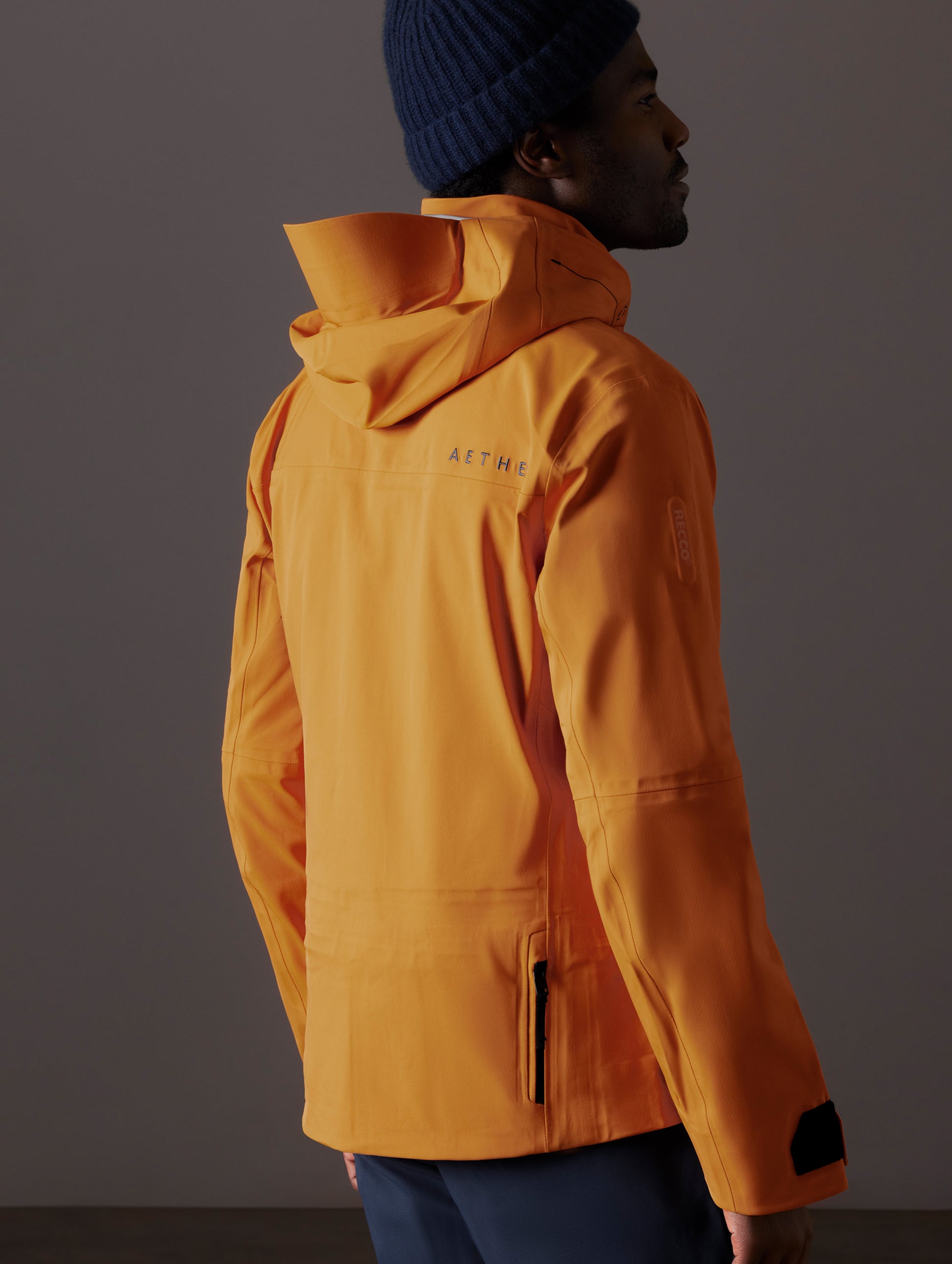 Rear 3/4 body view of man wearing Altitude Snow Shell 2.0 in Autumn Orange from AETHER Apparel.