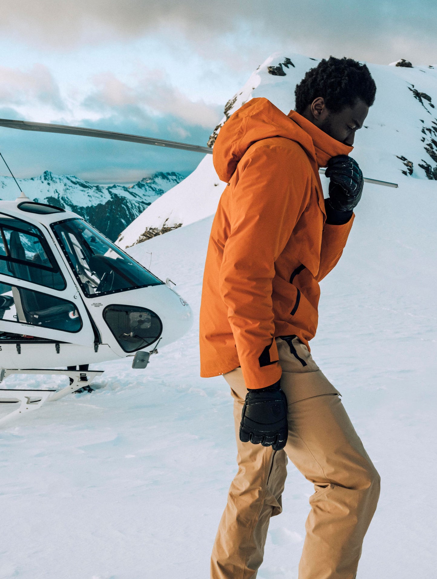 Man standing on snowy mountain top in front of helicopter wearing the Brighton Insulated Snow Jacket in Autumn Orange from AETHER Apparel.