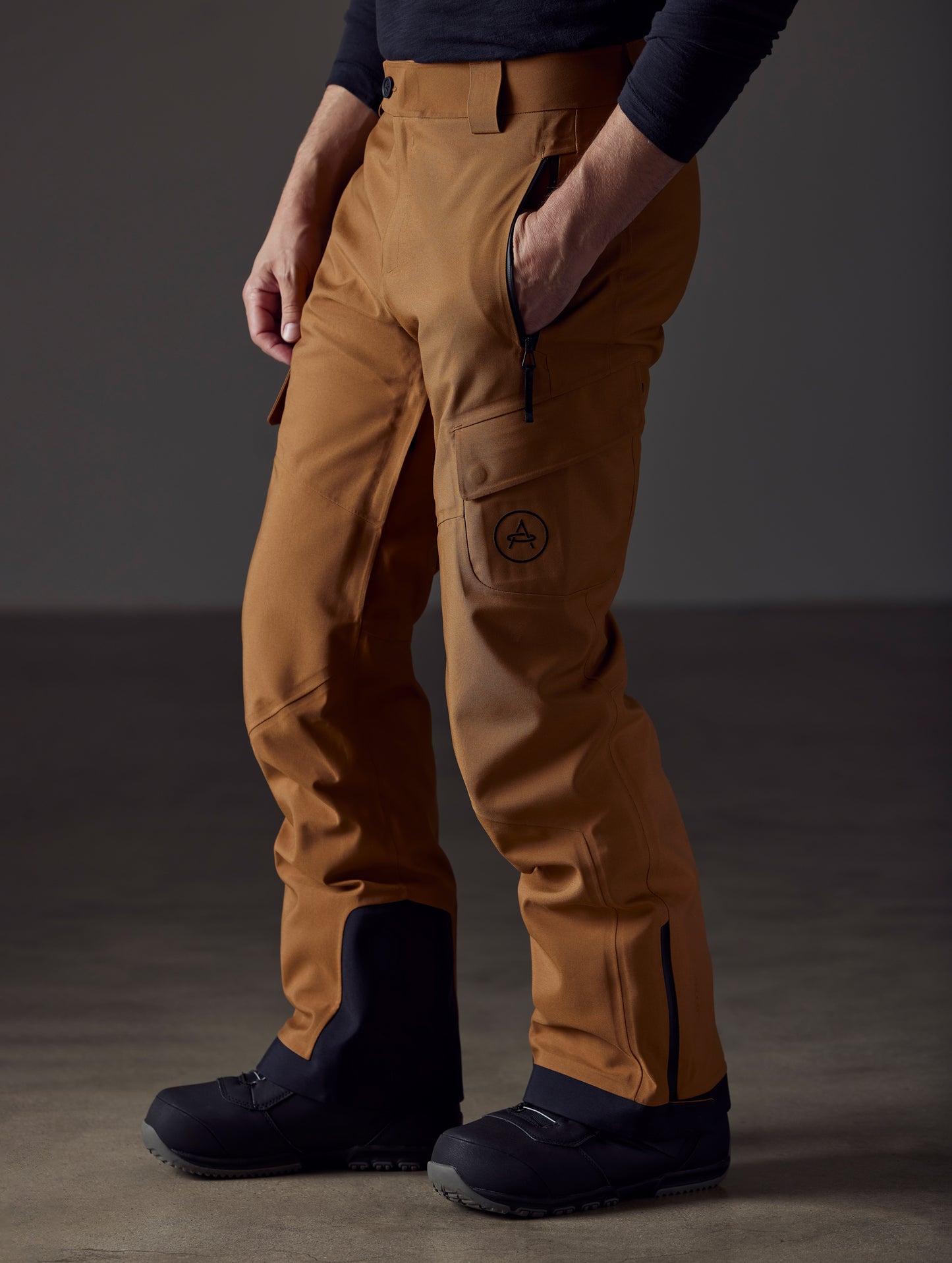 Front angled lower body view of man with knees bent and hand in pant pocket wearing Explorer Snow Pant in Rubber Brown from AETHER Apparel.