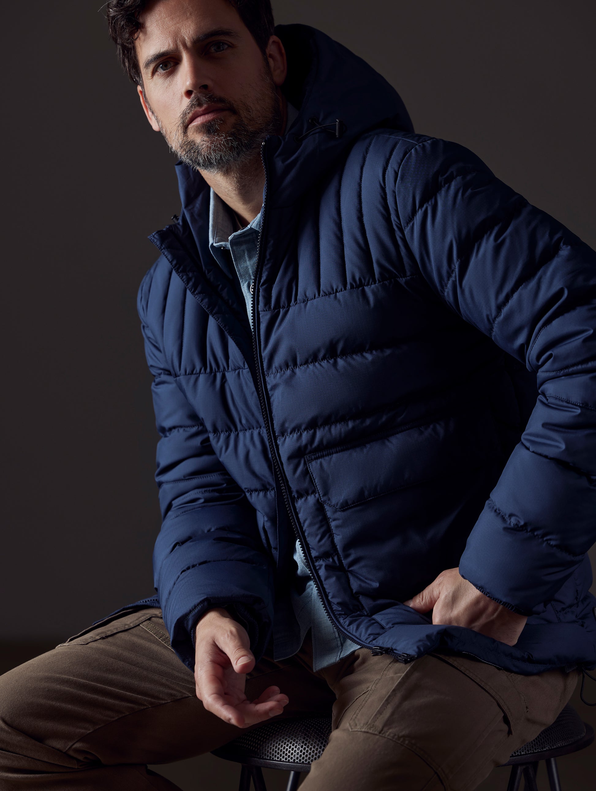 Front 3/4 body view of man sitting wearing Hudson Down Jacket in Total Eclipse dark blue from AETHER Apparel.