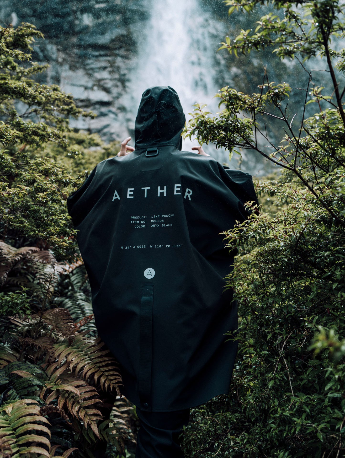 Outdoor lifestyle view of back of man walking through jungle in front of waterfall wearing the Line Poncho from AETHER Apparel.