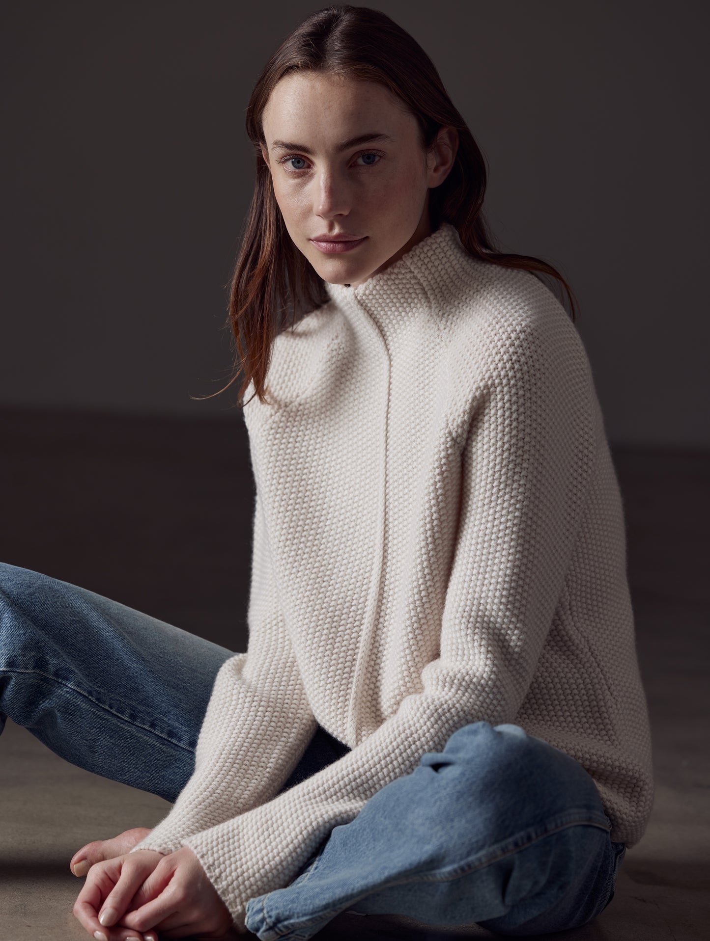 Front angled view of woman sitting on floor wearing jeans and Luxe Seamed Sweater in Winter White from AETHER Apparel.