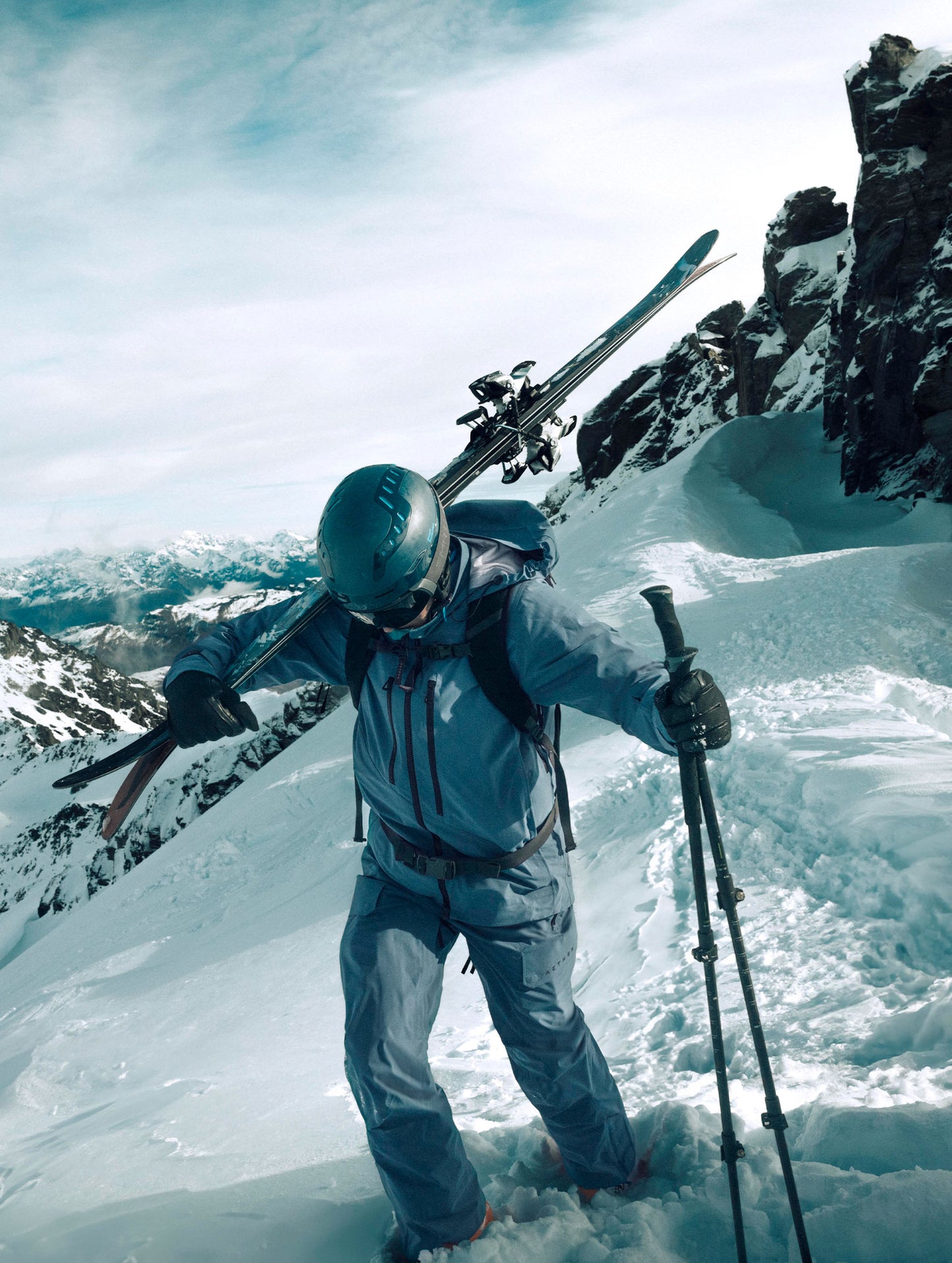 Man with skis over shoulder and poles in hand walking through snow on a mountain top wearing the Maverick Snow-Shell Jacket in Portal Blue from AETHER Apparel.