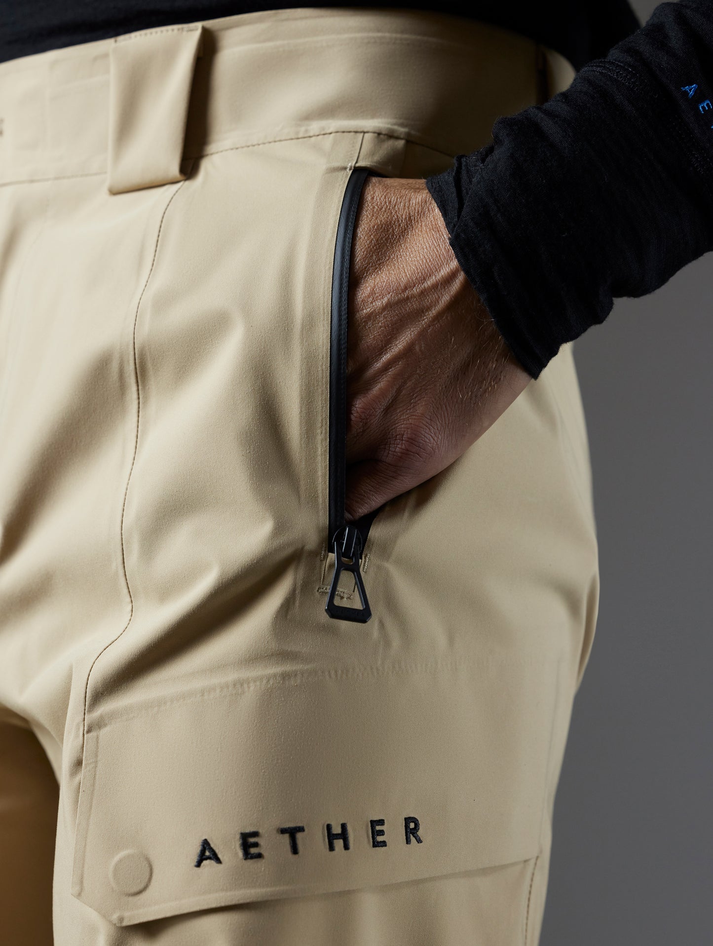 Closeup detailed view of the weatherproof zipper-secured, soft-lined hand pockets and and snap-secured, flap-covered cargo pockets on the Maverick Snow-Shell Pant in Desert Tan from AETHER Apparel.