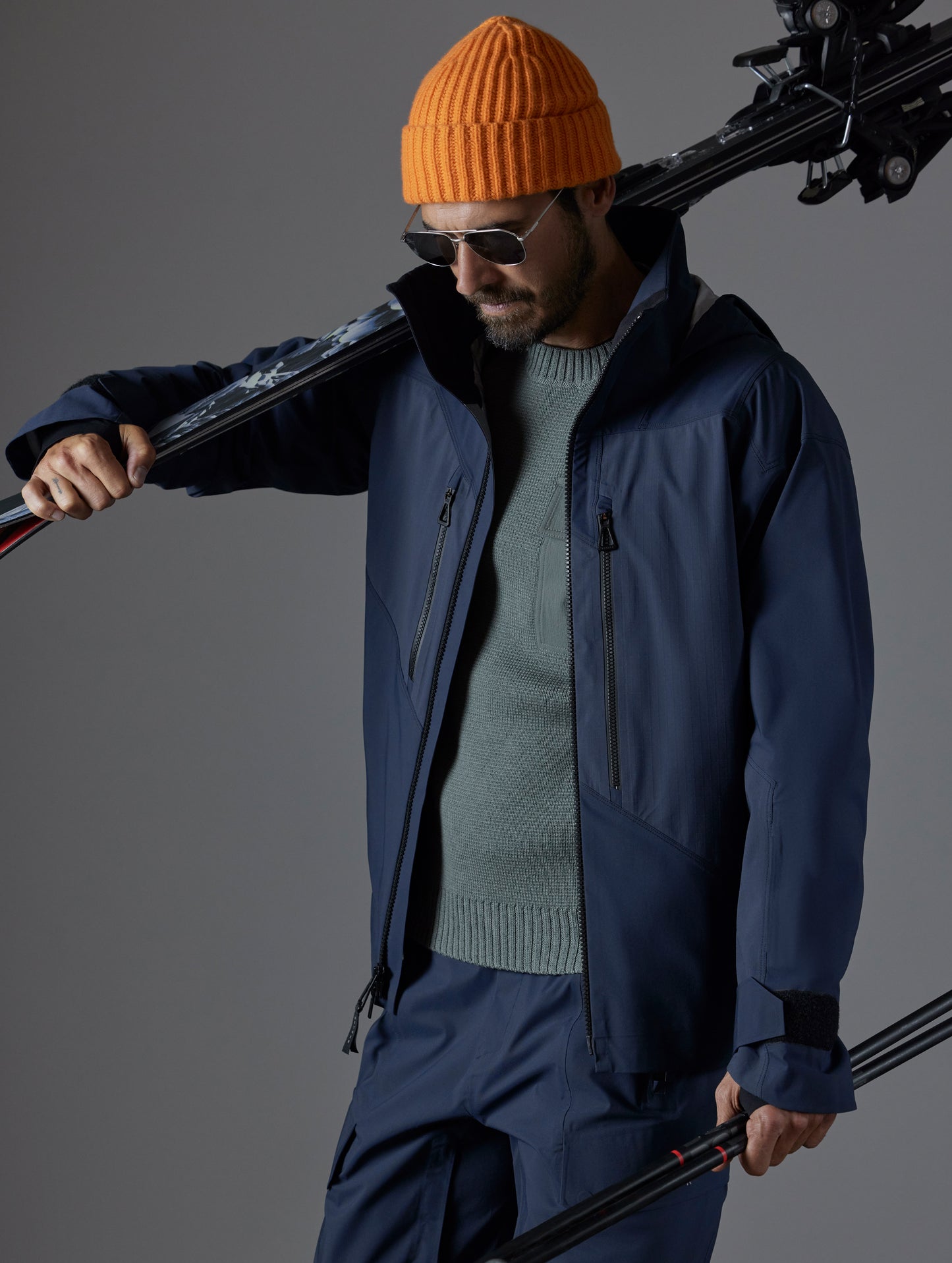 Front 3/4 full-body view of man holding skis over shoulder wearing Maverick Snow-Shell Jacket in Total Eclipse dark blue from AETHER Apparel.