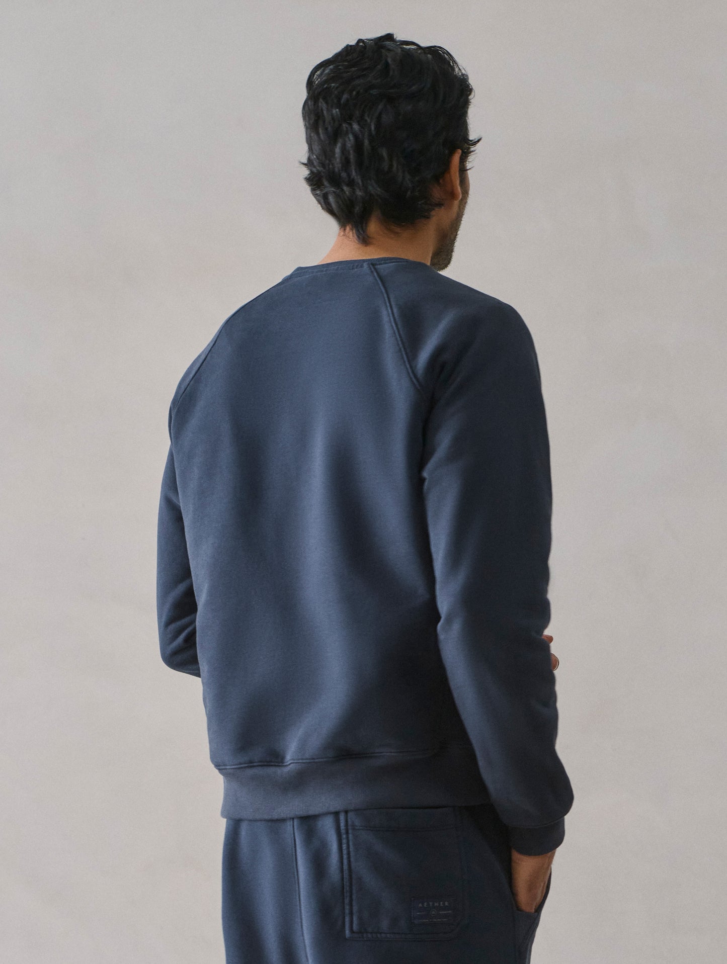 Rear 3/4 upper body view of man wearing Osa Raglan Sweatshirt in Total Eclipse dark blue from AETHER Apparel.