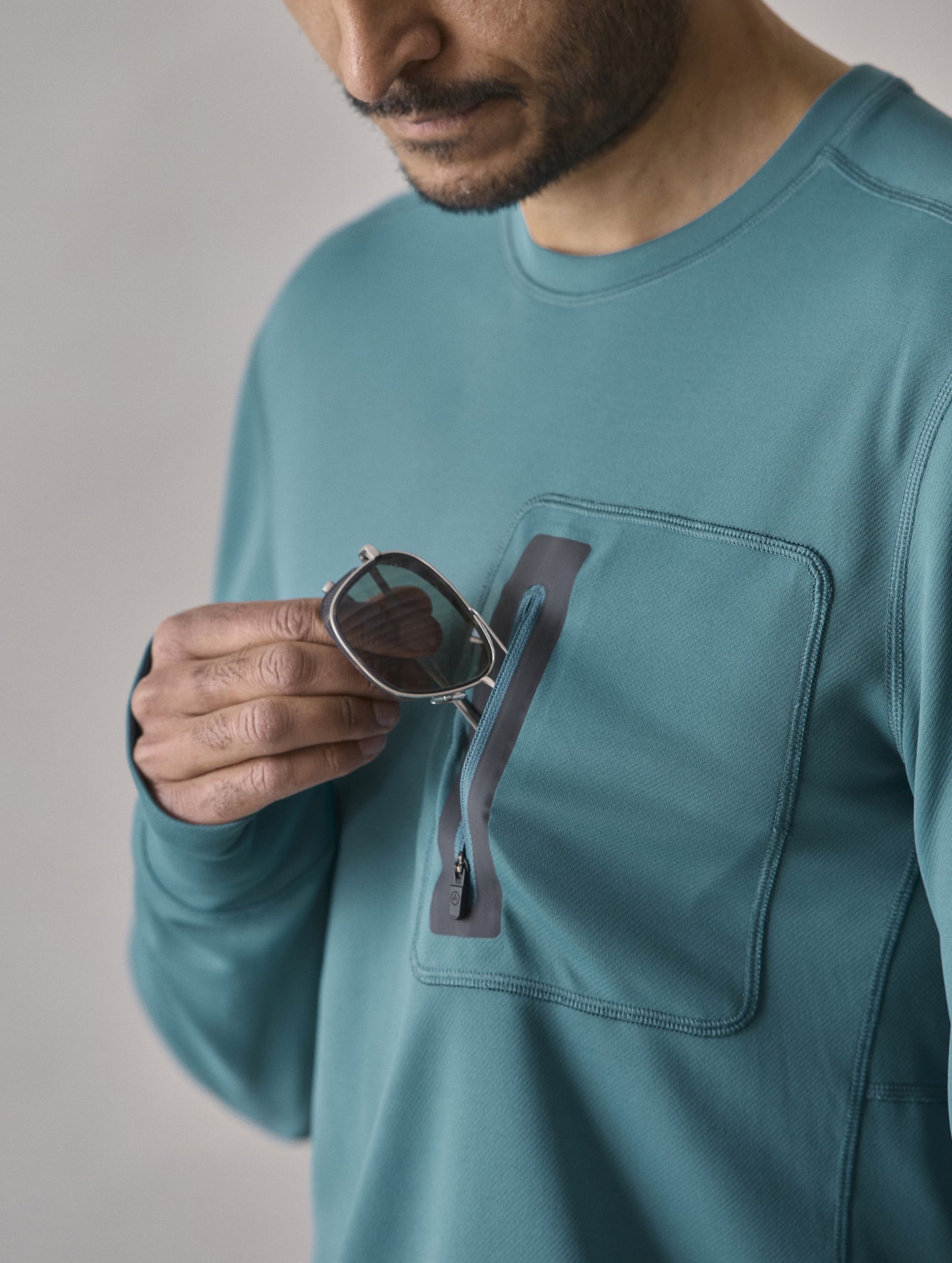 Closeup detailed view of man removing AETHER Summit Sunglass from the zipper-secured offset left chest patch pocket on the Pacific Crewneck Sun Shirt in Aqua Blue from AETHER Apparel.