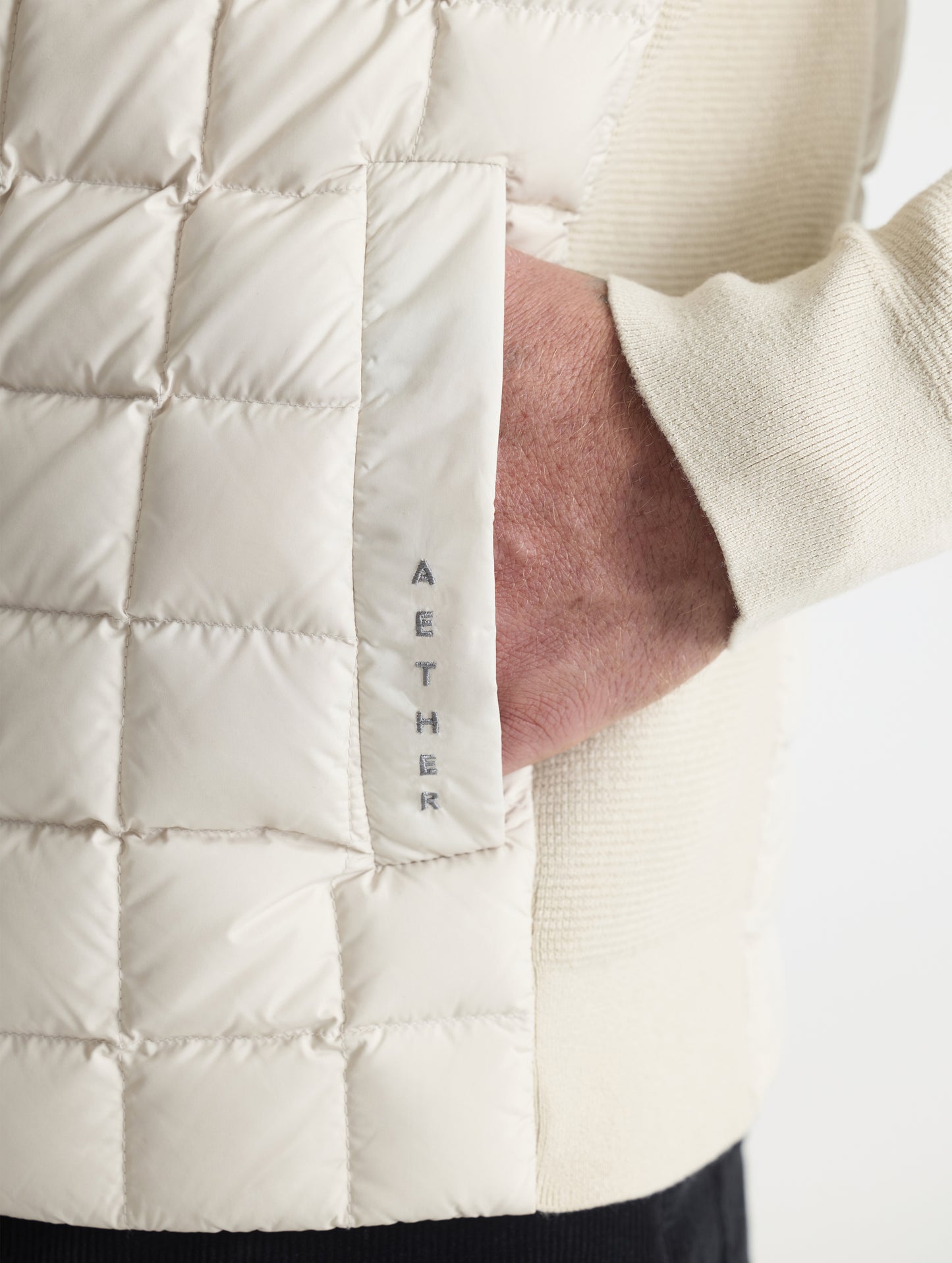Closeup detailed view of the embroidered AETHER logo and welt hand pocket on the M Phase Full-Zip in Bone white from AETHER Apparel.