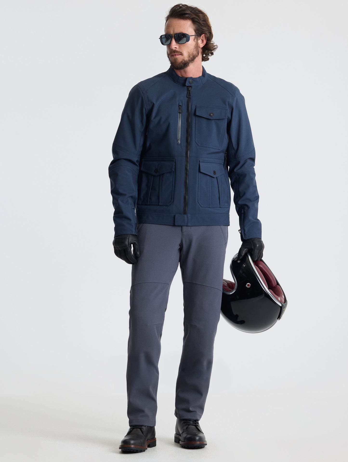 Front full-body view of man holding motorcycle helmet wearing Ramble Pant 3.0 in Graphite from AETHER Apparel.