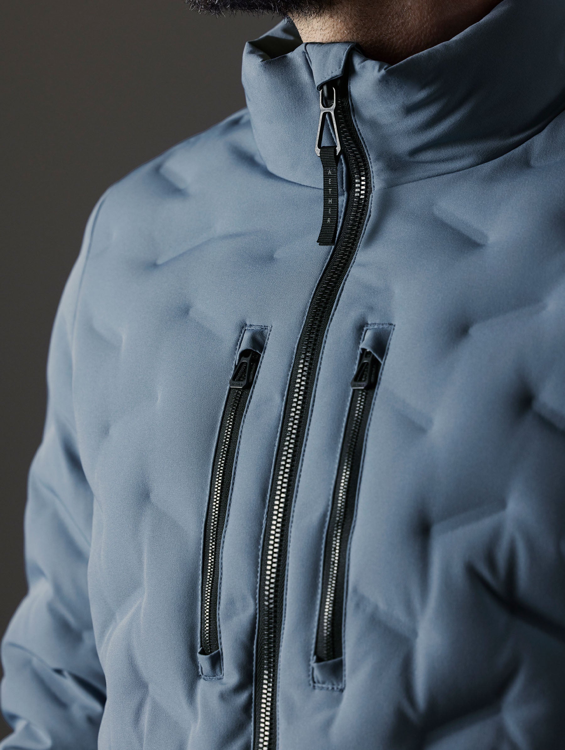 Closeup detailed view of the main center-front zipper and zippered chest pockets on the Rex Down Jacket in Portal Blue from AETHER Apparel.