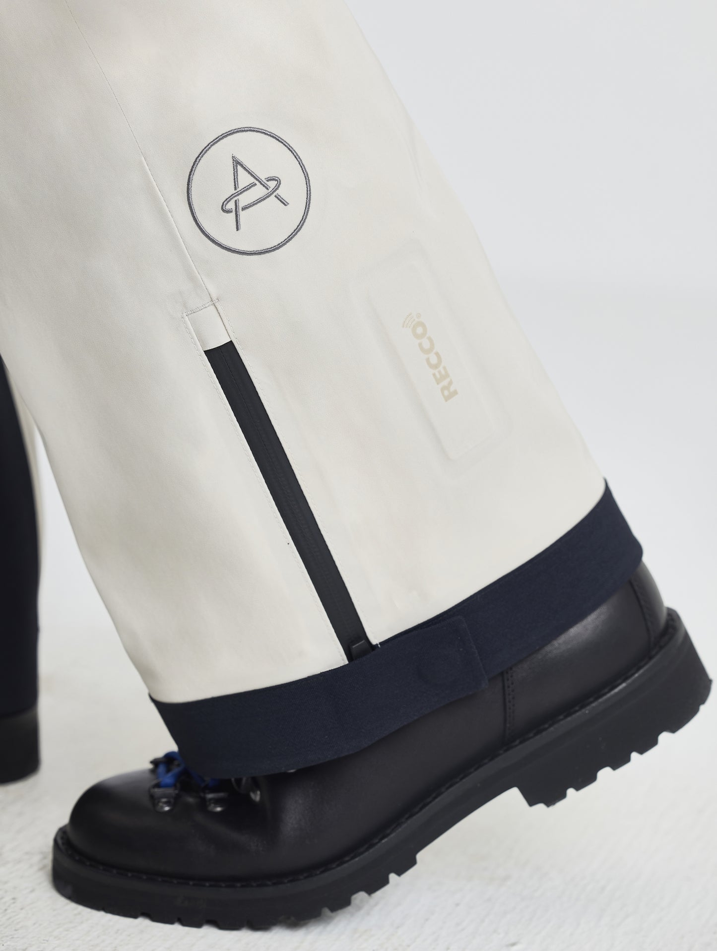 Closeup detailed view of the AETHER logo and zipper-adjustable leg cuffs on the Ridge Insulated Snow Pant in Bone from AETHER Apparel.