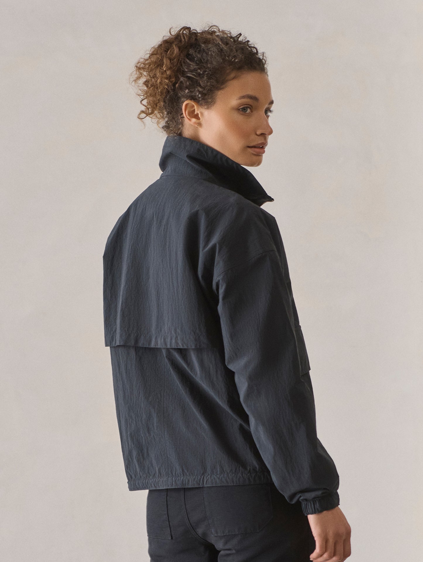 Rear 3/4 body view of woman wearing Mila Windbreaker in Onyx Black from AETHER Apparel.
