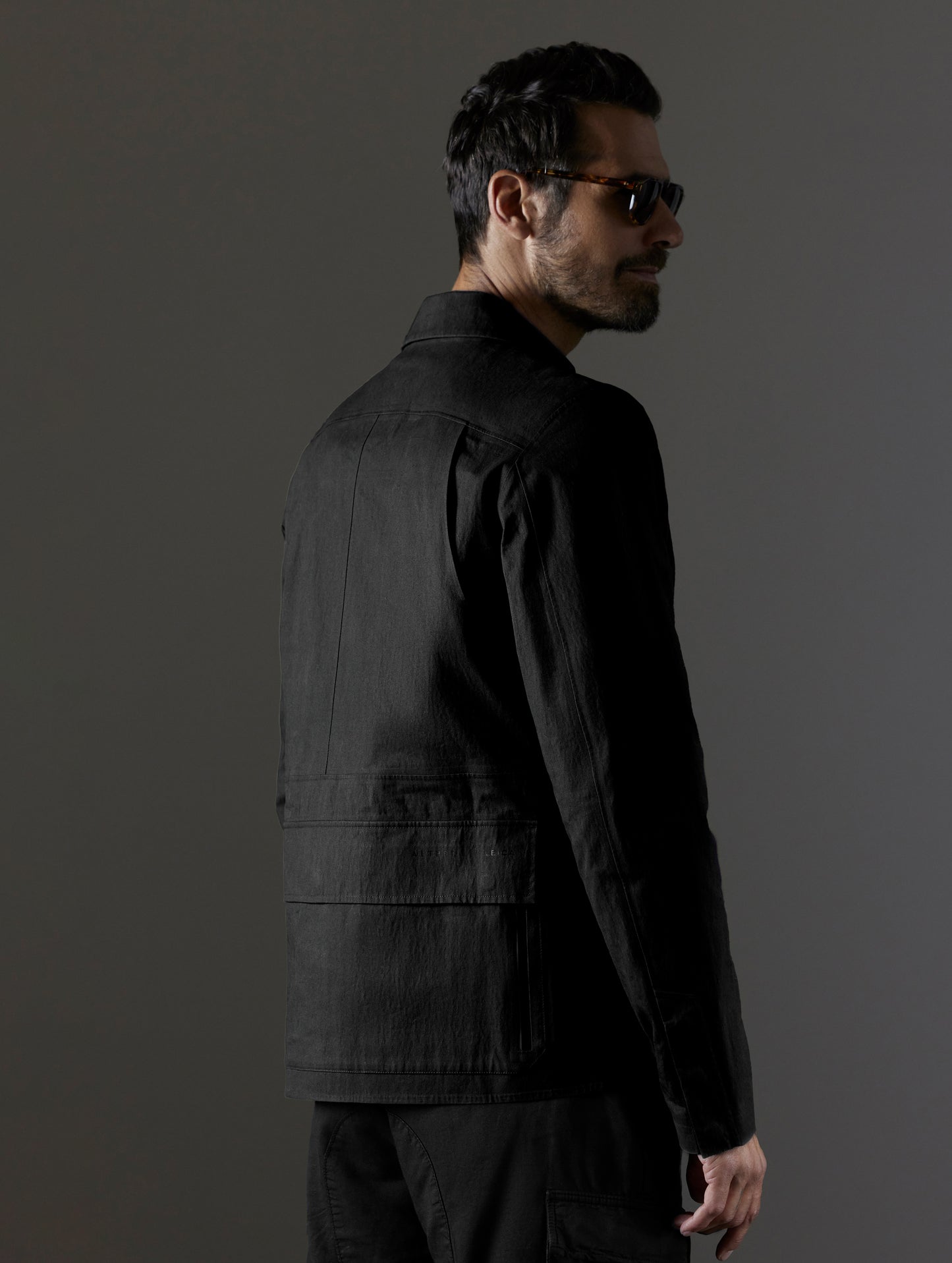 Rear 3/4 upper body view of man wearing buttoned-up AETHER + LEICA Reporter Jacket in Monochrom.