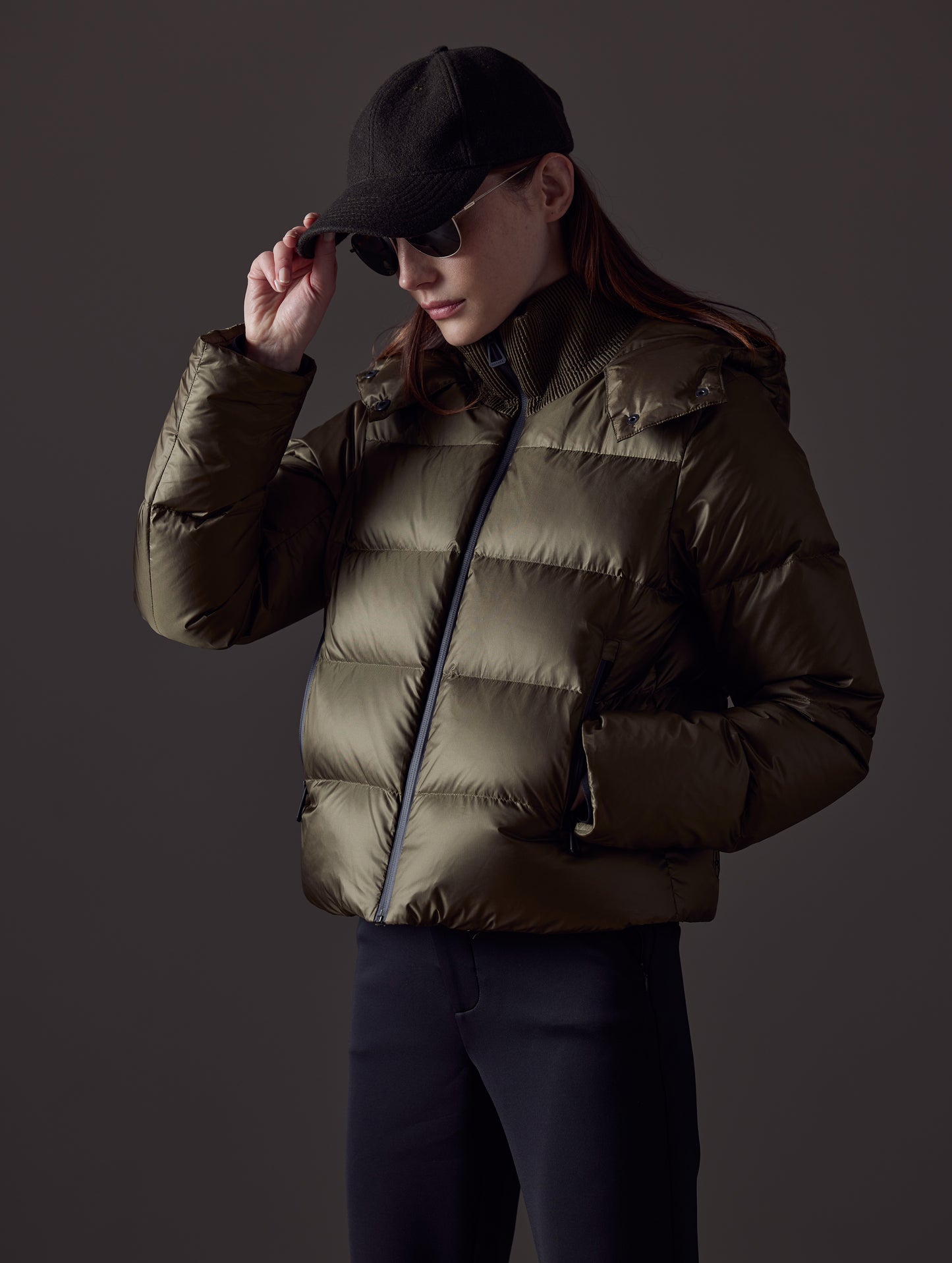 Front angled view of woman with hand on cap wearing Aura Puffer Jacket in Command Green from AETHER Apparel.