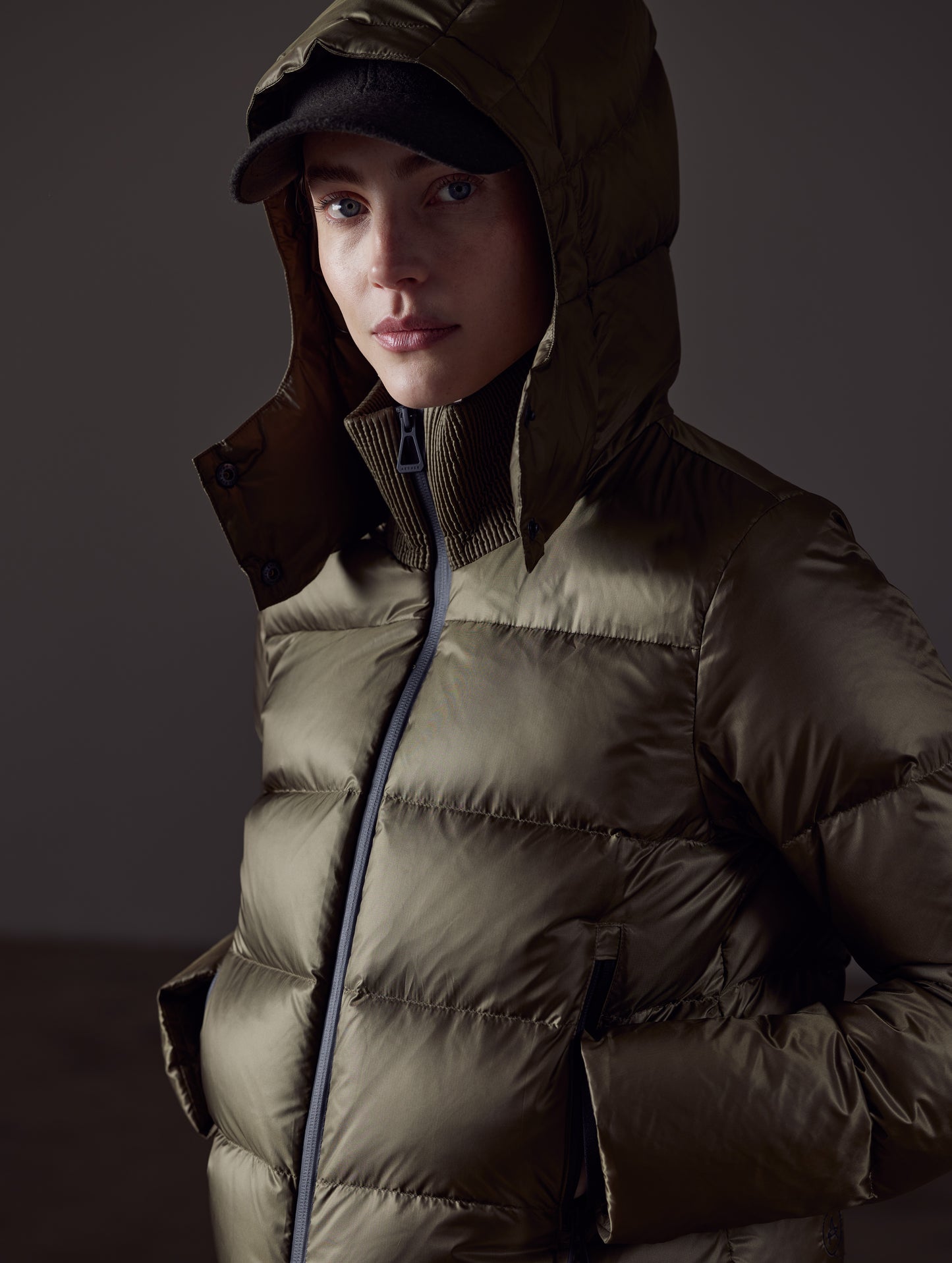 Closeup detailed front angled view of woman with hood up wearing Aura Puffer Jacket in Command Green from AETHER Apparel.