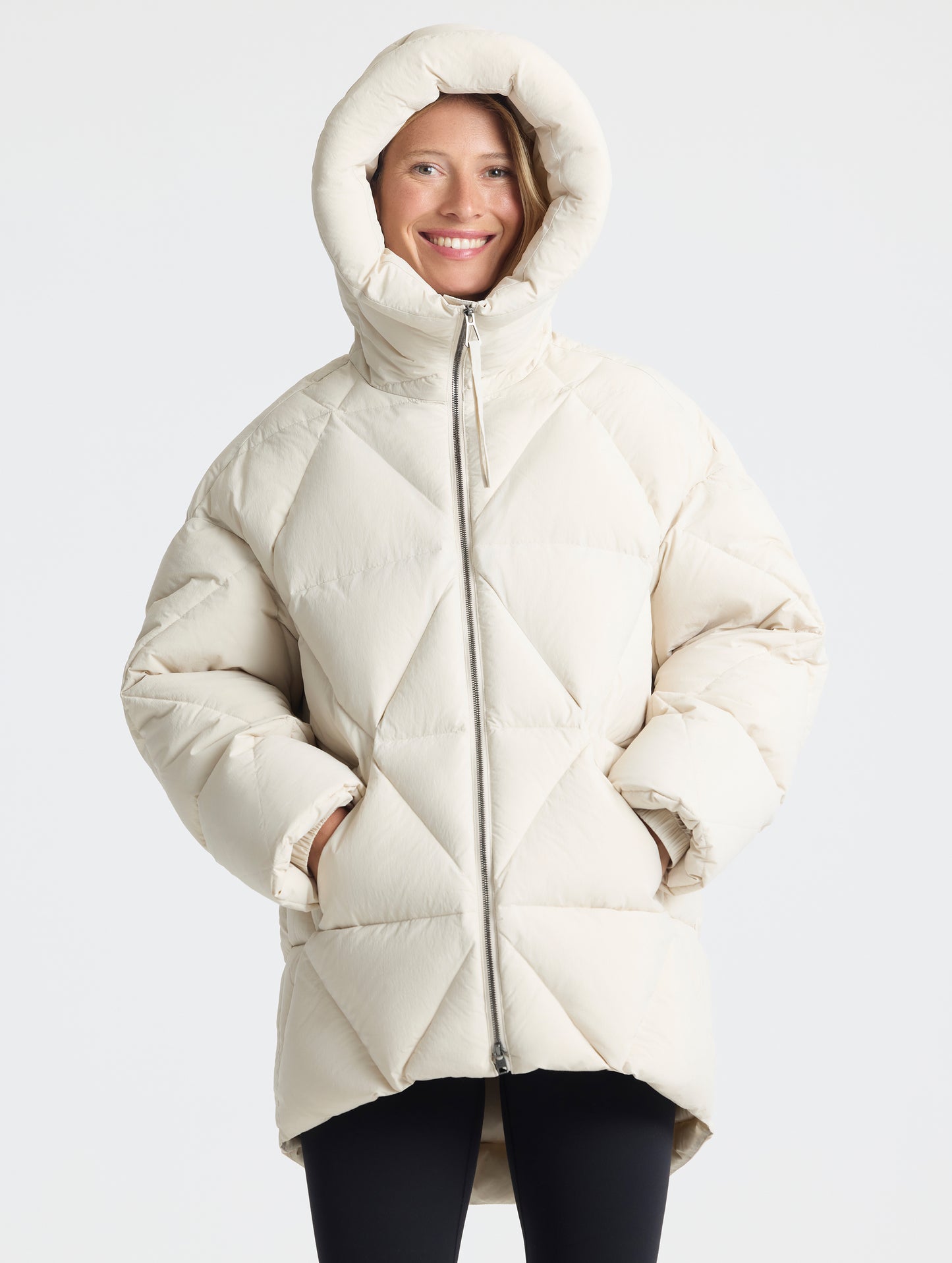 Front upper-body view of woman with hood up wearing a fully zipped up Callie Oversized Down Parka in Bone from AETHER Apparel.
