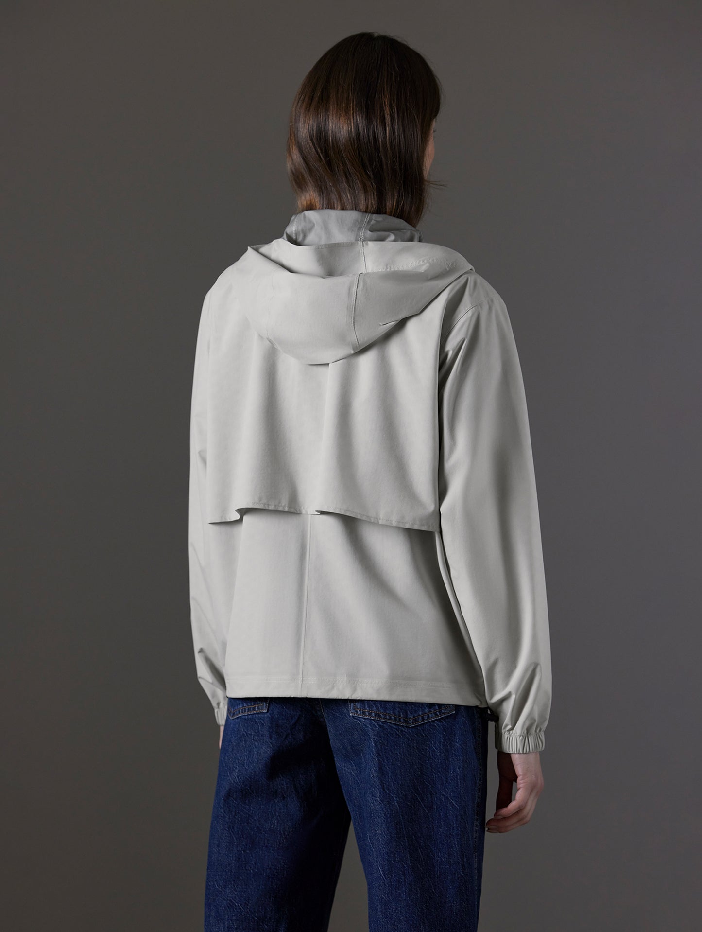 Rear angled body view of woman wearing Kite Lightweight Jacket in Stone Grey from AETHER Apparel.