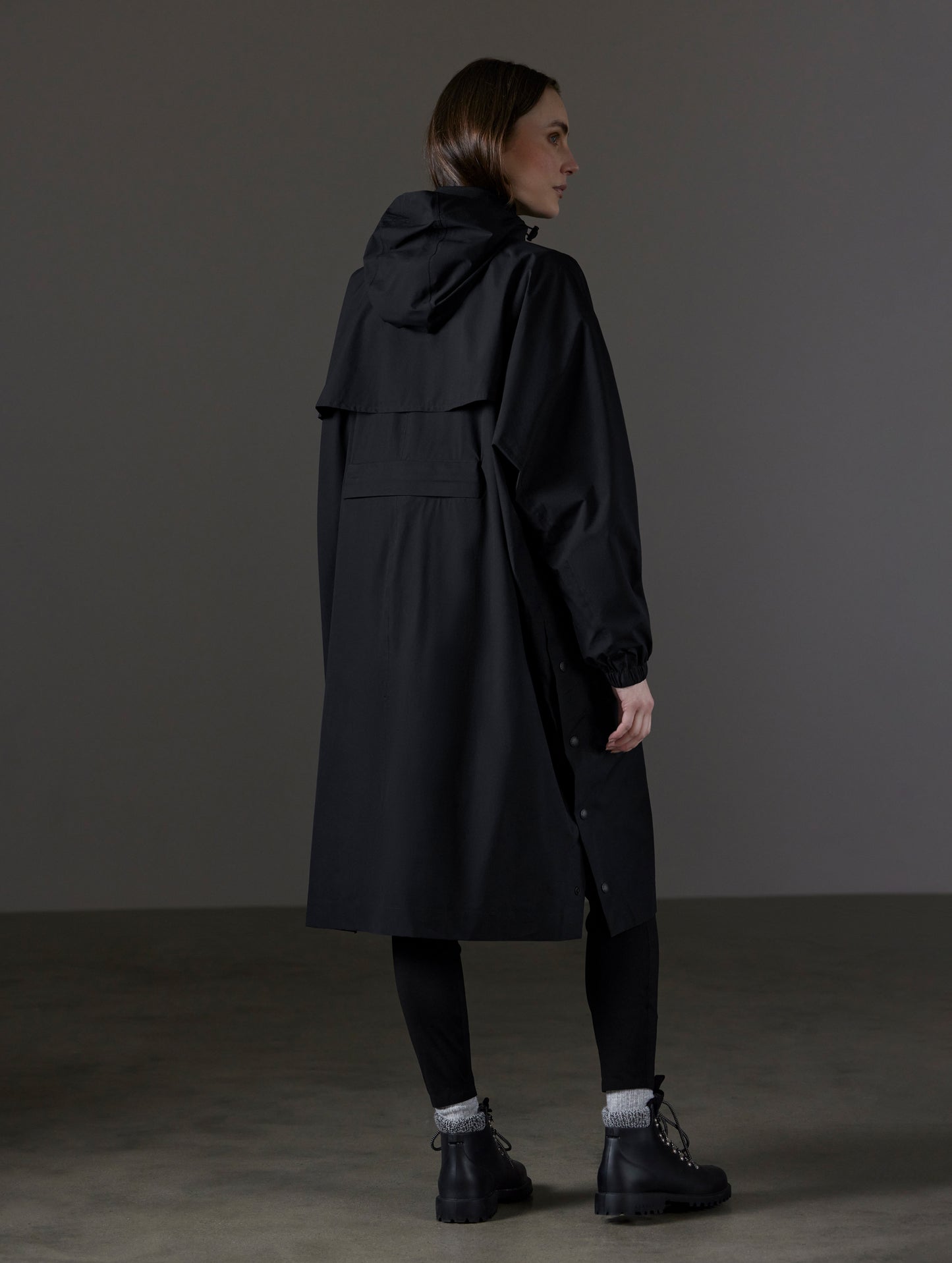 Rear angled full-body view of woman wearing Kite Packable Rain Poncho in Onyx Black from AETHER Apparel.