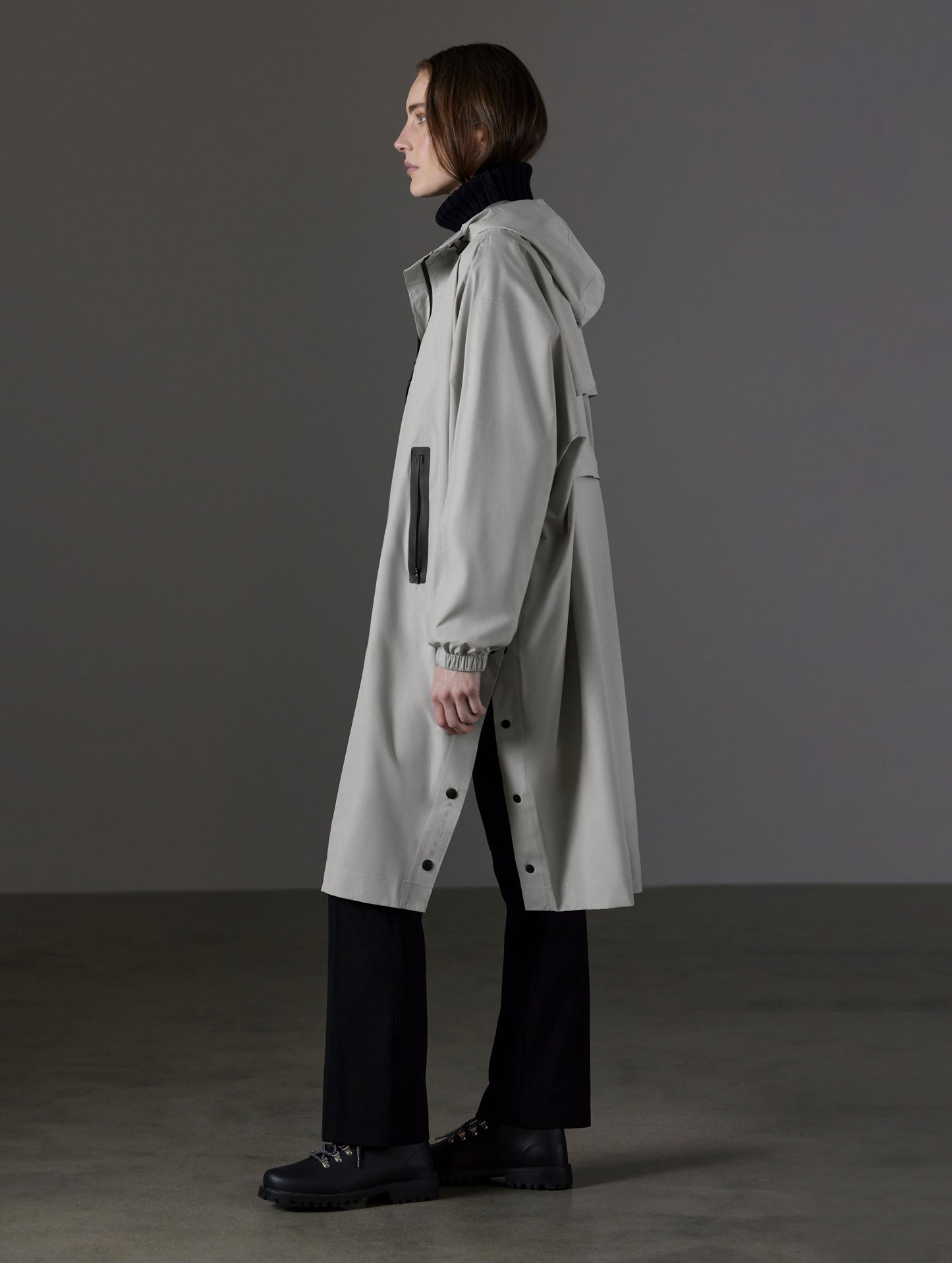 Side profile full-body view of woman wearing Kite Packable Rain Poncho in Stone Grey from AETHER Apparel.