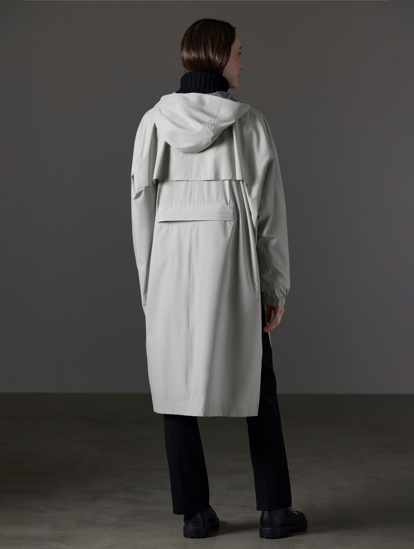 Rear angled full-body view of woman wearing Kite Packable Rain Poncho in Stone Grey from AETHER Apparel.
