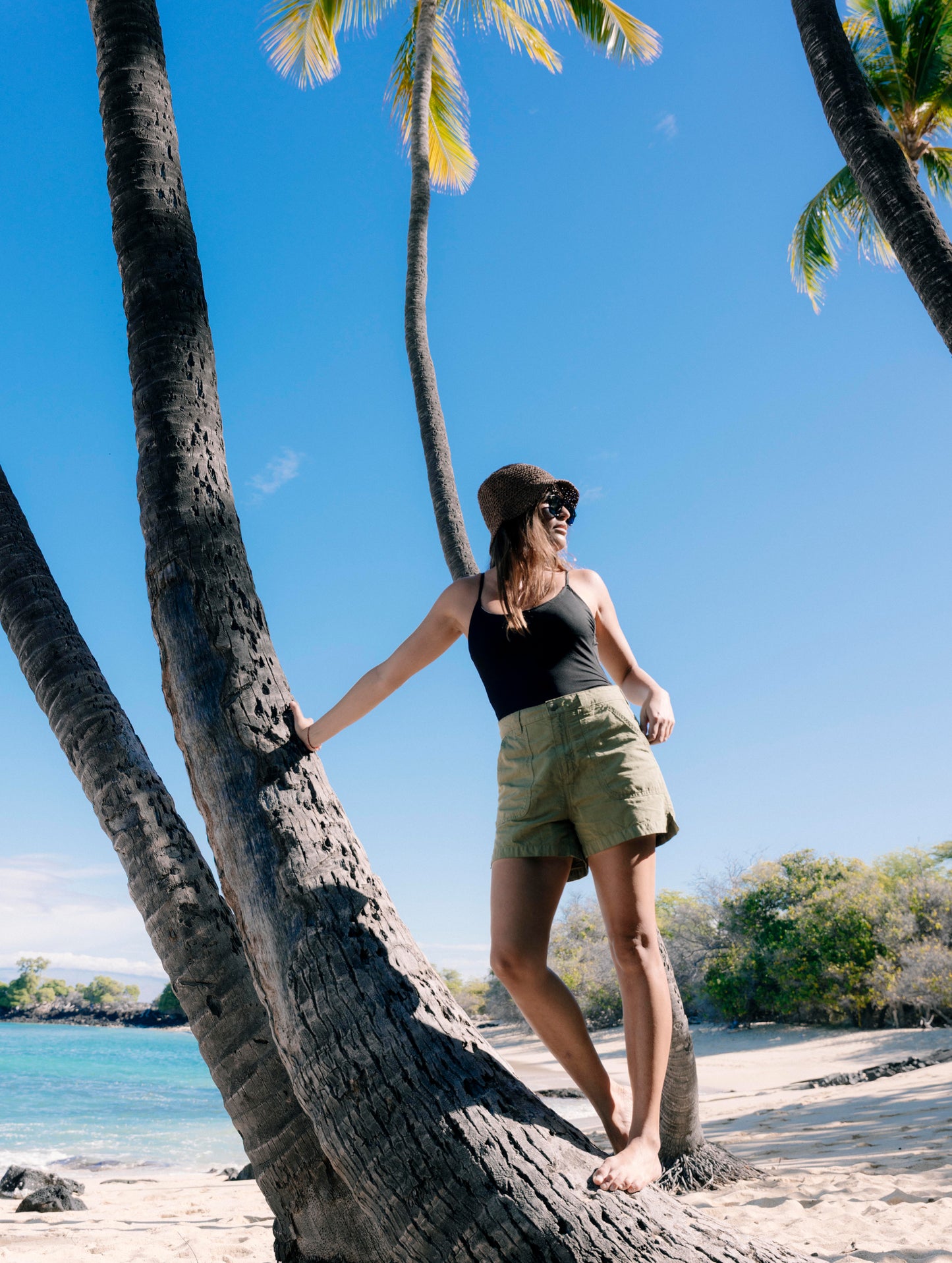 Lifestyle image of woman standing and leaning against a palm tree while wearing the Masai Safari Short in Fern Green from AETHER Apparel.