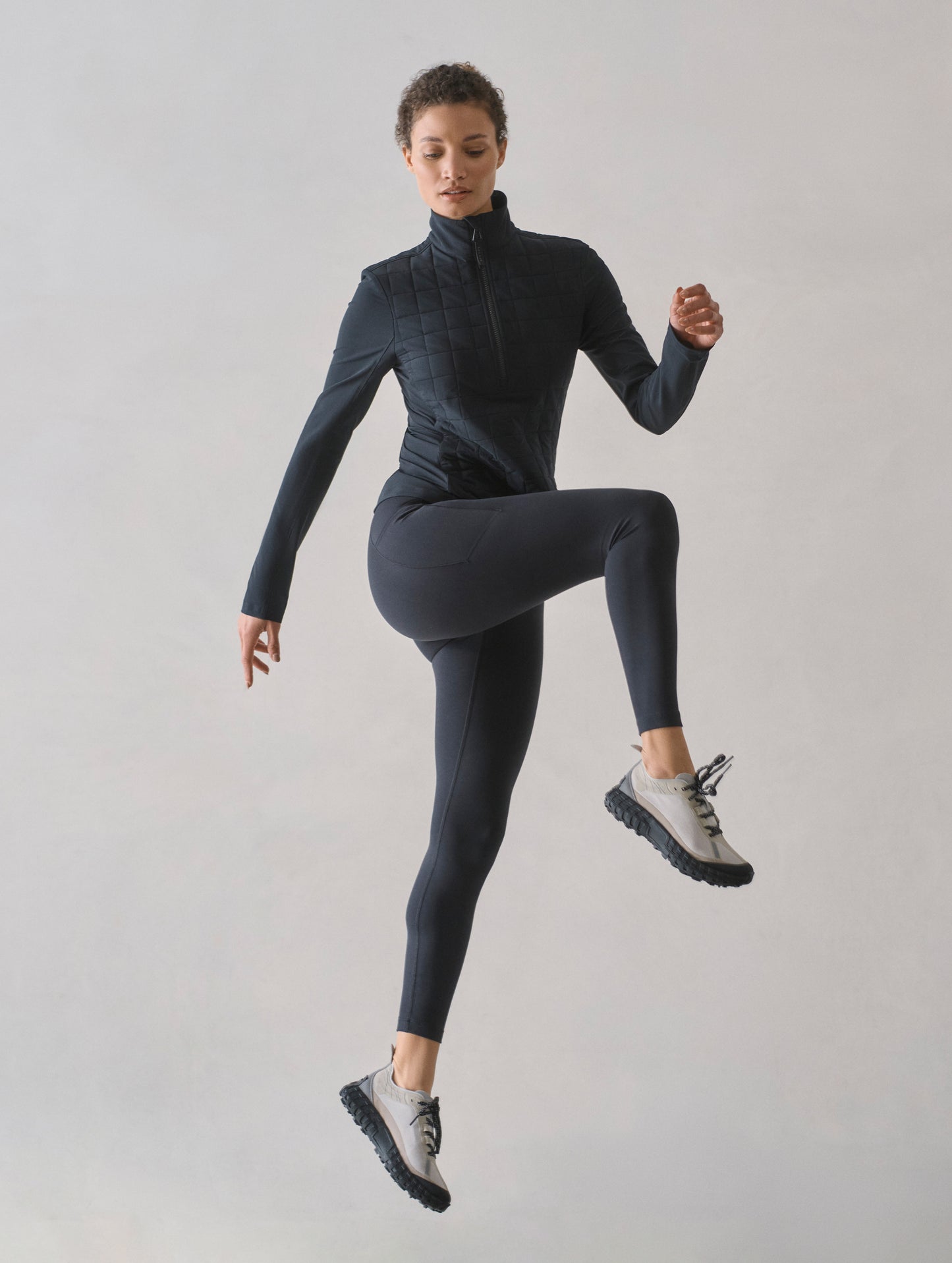 Studio image of woman jumping while wearing Wren Insulated Half-Zip in Onyx Black from AETHER Apparel.
