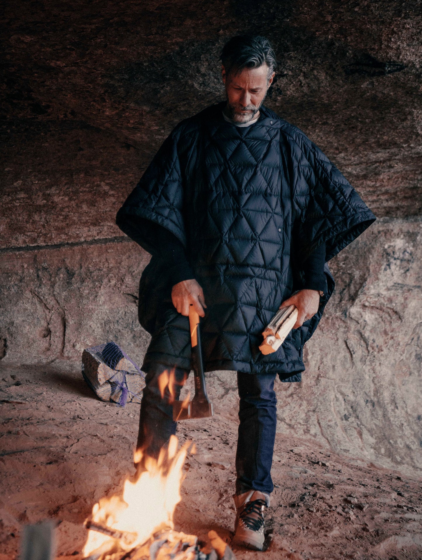 Outdoor lifestyle image of man standing over campfire wearing Down Poncho in Onyx Black from AETHER Apparel.