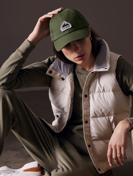 Front angled view of woman sitting on floor wearing AETHER Mountain Hat in Army Green from AETHER Apparel.