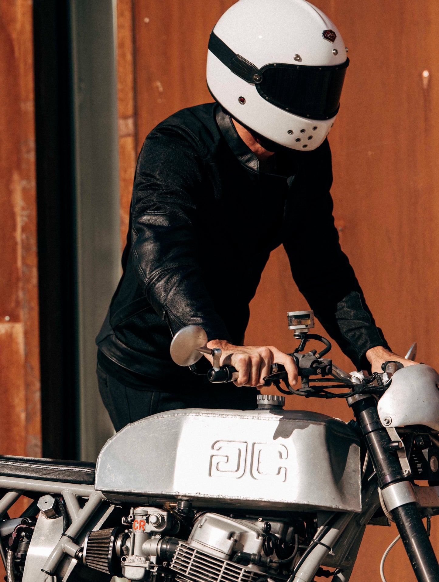 Outdoor lifestyle image of man pushing custom motorcycle wearing white helmet and Draft Mesh Motorcycle Jacket in Jet Black from AETHER Apparel.