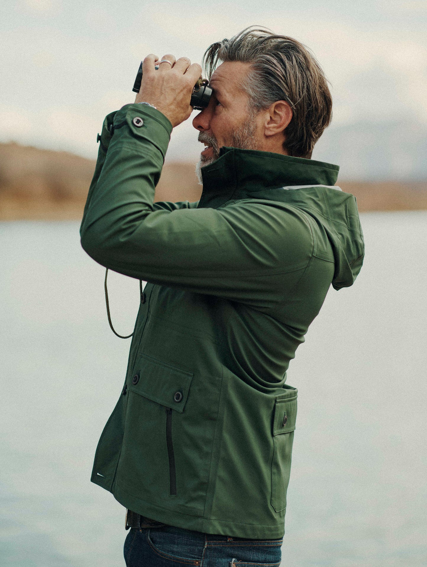 Outdoor lifestyle image of man looking through binoculars wearing Endeavor Jacket in Duffle Green from AETHER Apparel.