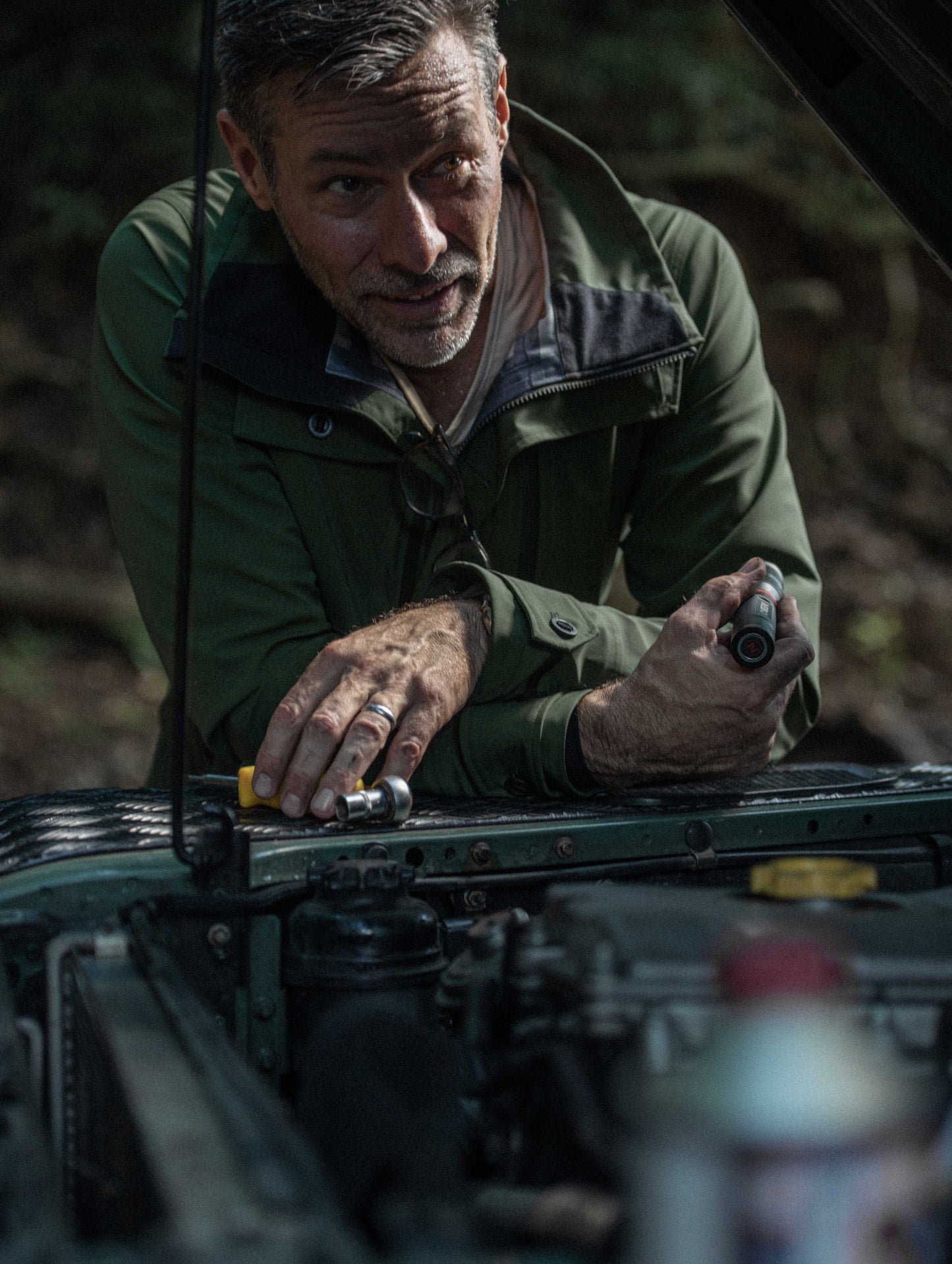 Outdoor lifestyle image of man leaning over open hood of Land Rover Defender wearing Endeavor Jacket in Duffle Green from AETHER Apparel.