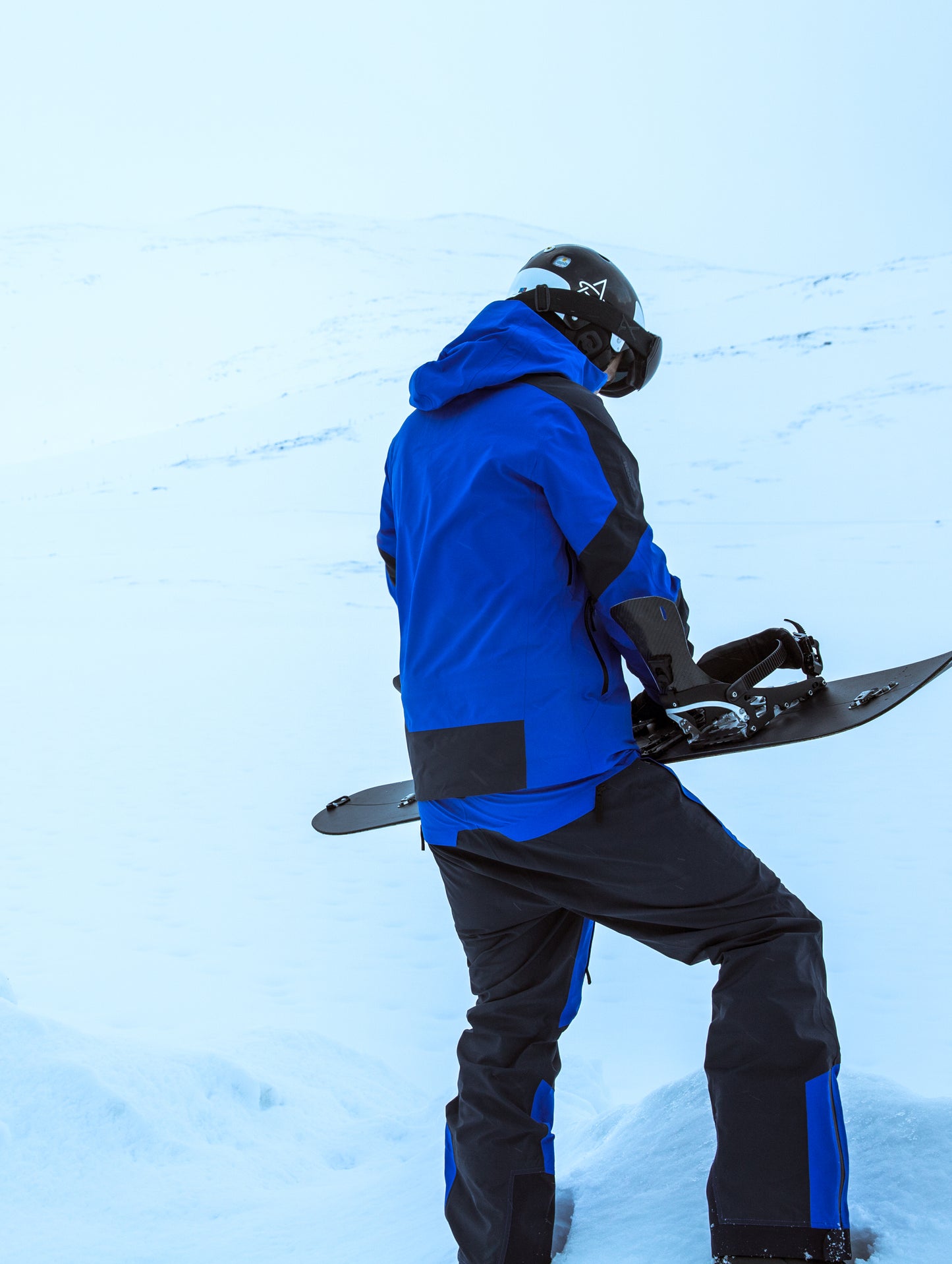 Man outdoors holding snowboard wearing blue Fusion Shell from AETHER Apparel.