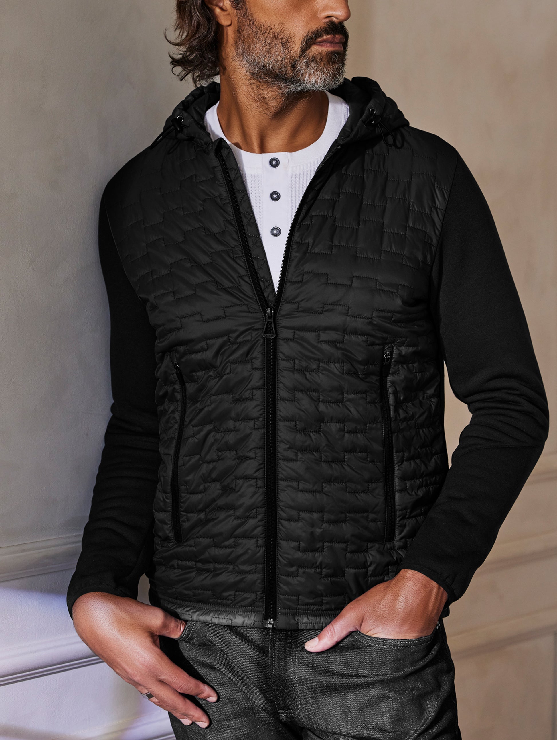 Man standing indoors wearing insulated Hybrid Tech Hoodie from AETHER Apparel.
