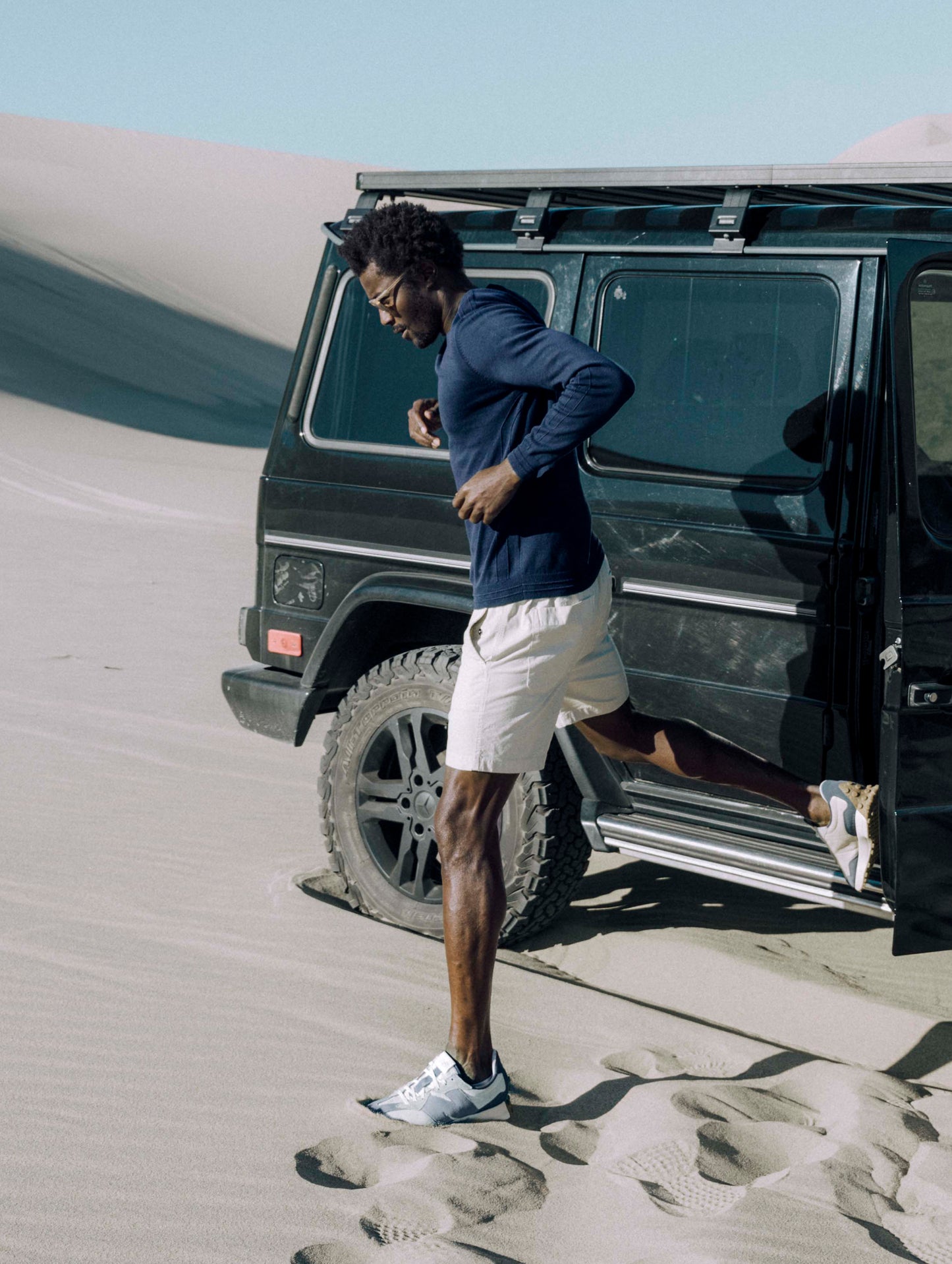 Outdoor lifestyle image of man standing on sand dunes next to SUV wearing Irving Sweater in Total Eclipse dark blue from AETHER Apparel.