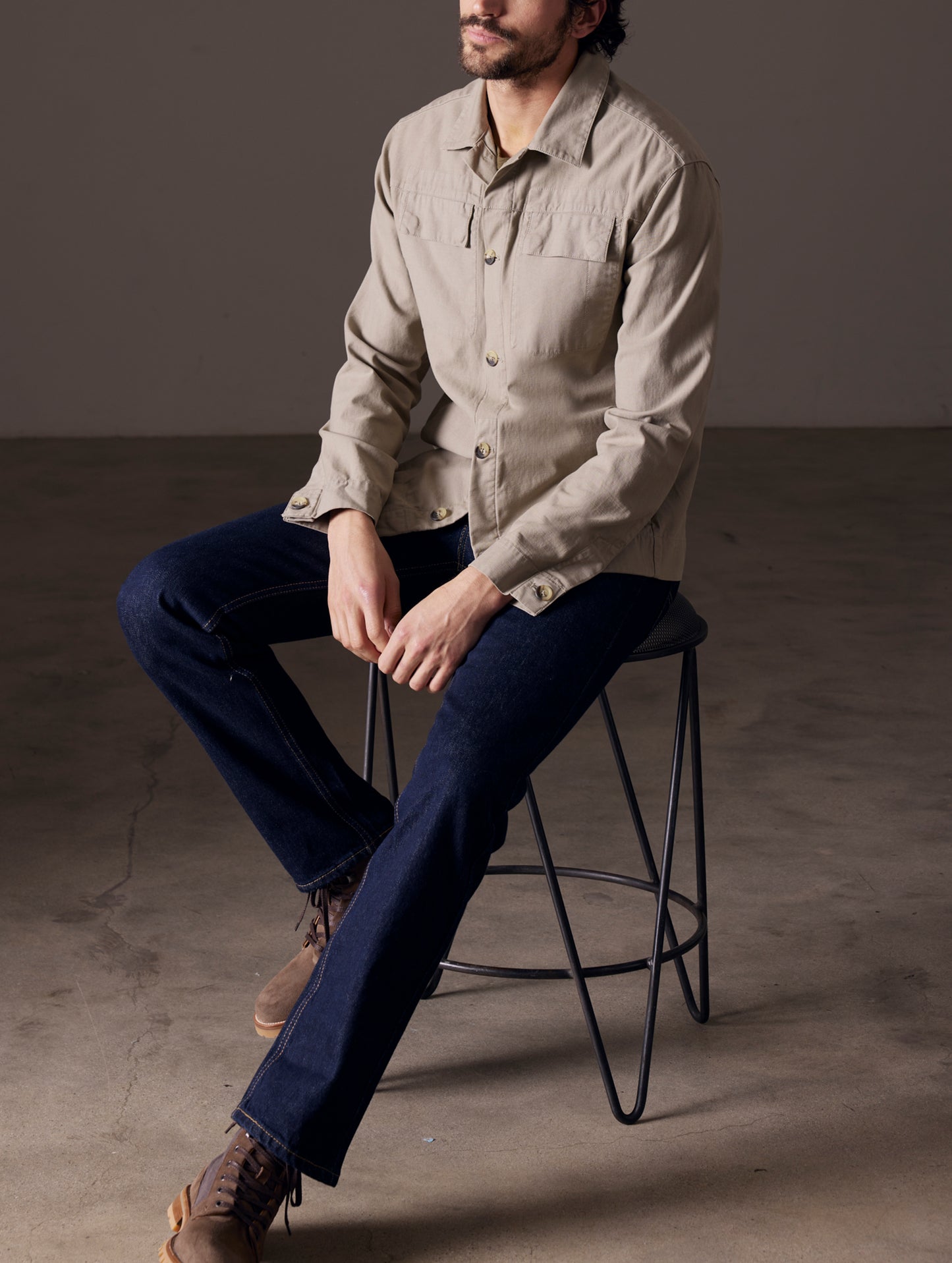 Front 3/4 full-body view of man sitting on stool wearing Mesa Jacket in Laurel Grey from AETHER Apparel.
