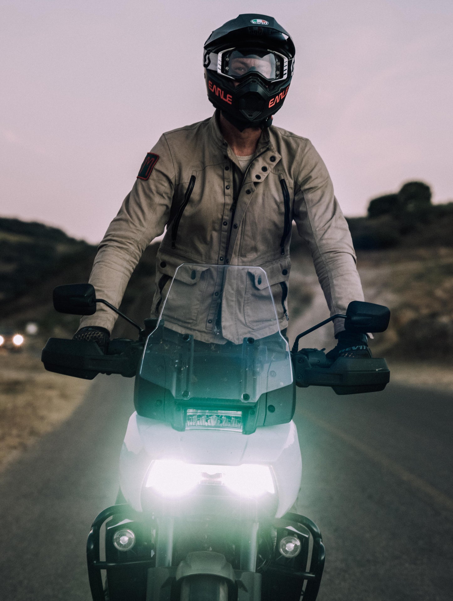 Outdoor lifestyle image of man standing up riding motorcycle wearing Mojave Motorcycle Jacket in Dark Sand from AETHER Apparel.