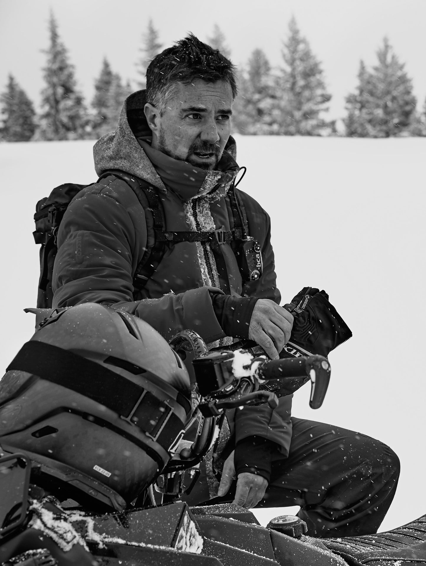 Black and white outdoor lifestyle image of man sitting on snowmobile wearing Nordic Jacket from AETHER Apparel.