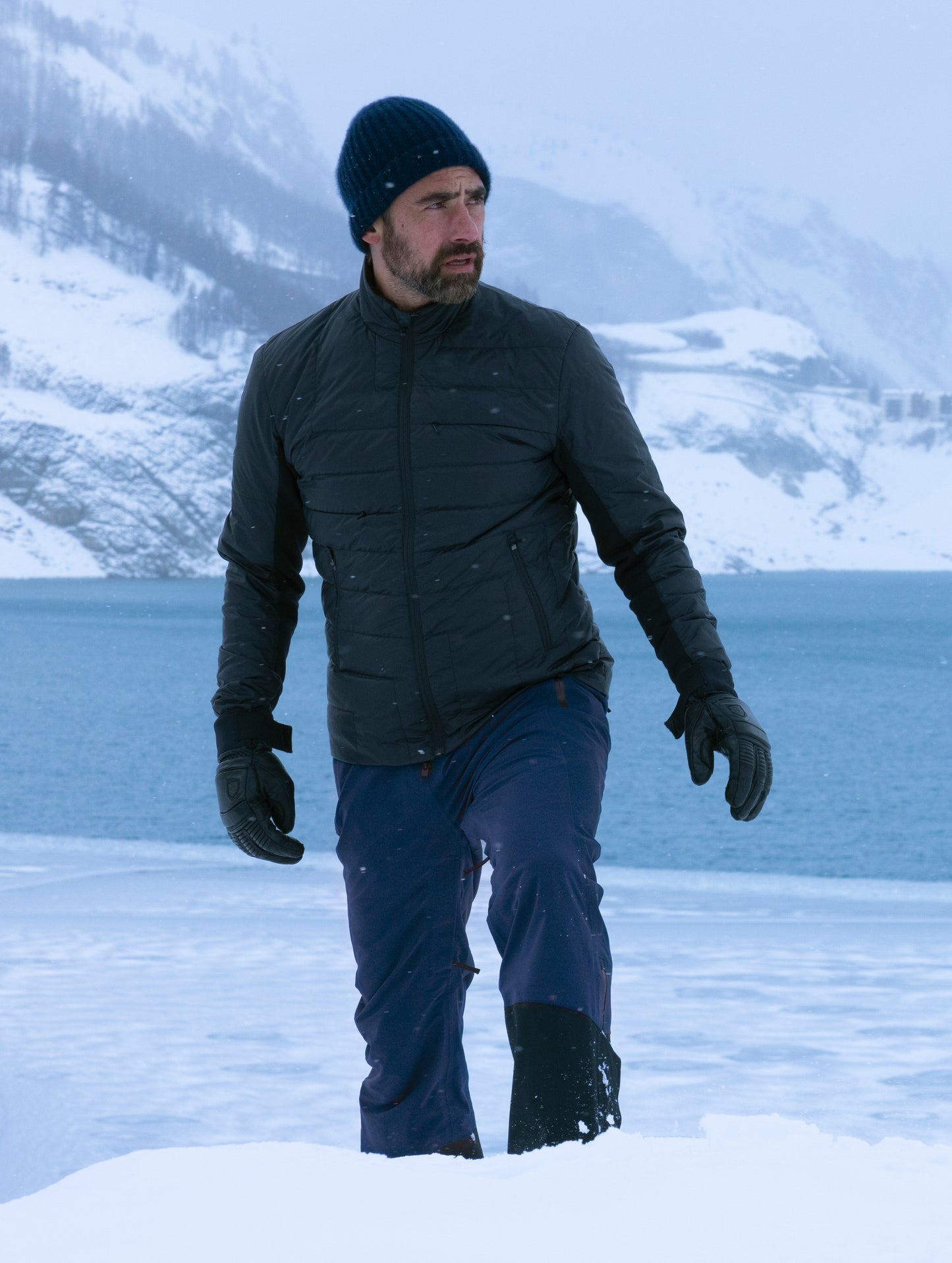 Outdoor lifestyle image of man standing in front of snowy mountains and lake wearing Stealth Snow Bib in Nordic Navy blue from AETHER Apparel.