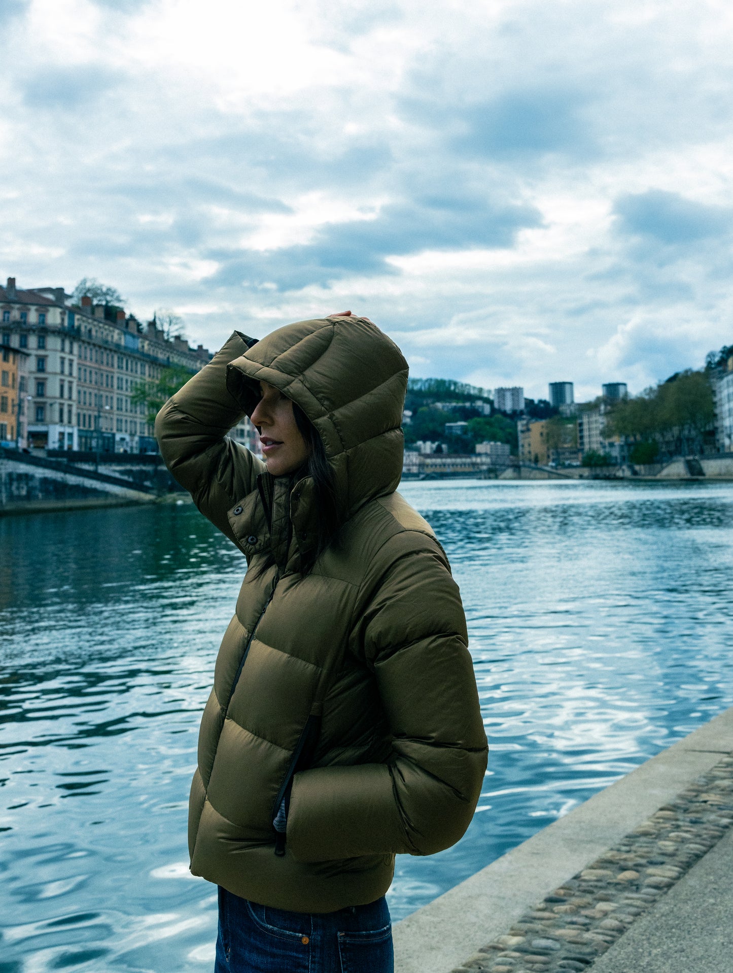 Outdoor lifestyle image of woman standing next to body of water wearing Aura Puffer Jacket in Command Green from AETHER Apparel.
