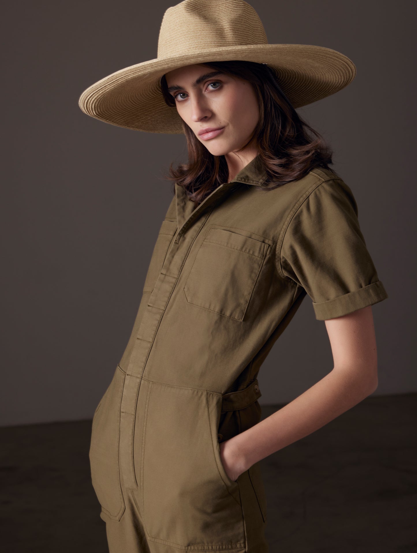 Front angled view of woman wearing sun hat and Bard Jumpsuit in Command Green from AETHER Apparel.