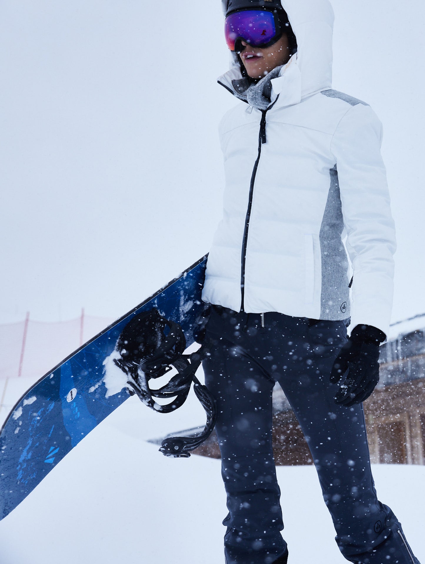 Outdoor lifestyle image of woman holding snowboard wearing Descent Snow Bib 2.0 in Total Eclipse dark blue from AETHER Apparel.