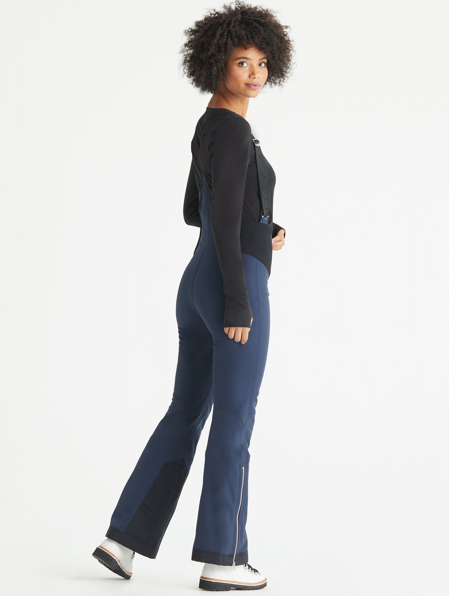Rear angled full-body view of woman wearing Descent Snow Bib 2.0 in Total Eclipse dark blue from AETHER Apparel.