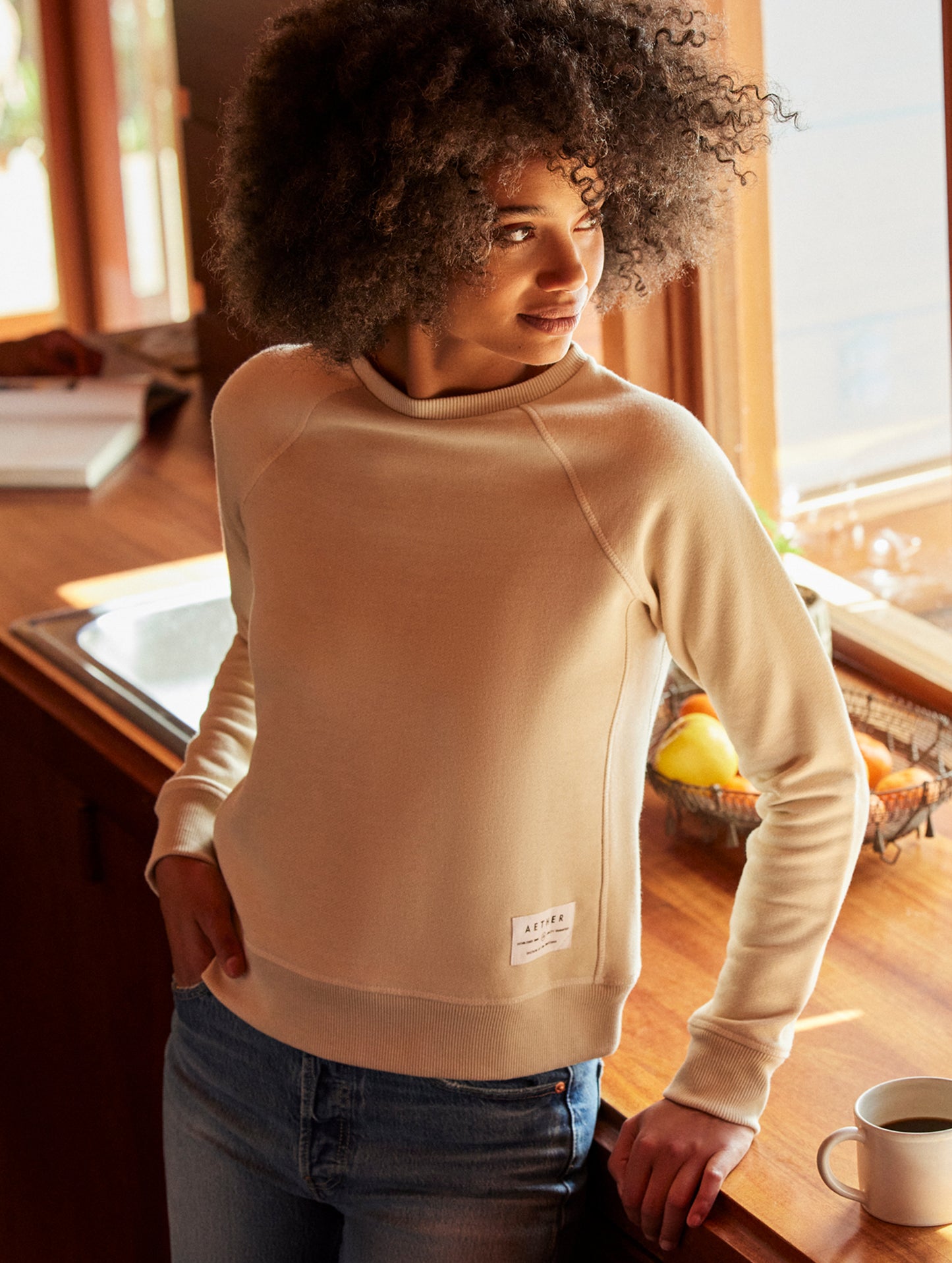 Front angled indoor lifestyle view of woman standing next to desk wearing Foundation Raglan Sweatshirt in Bone white from AETHER Apparel.