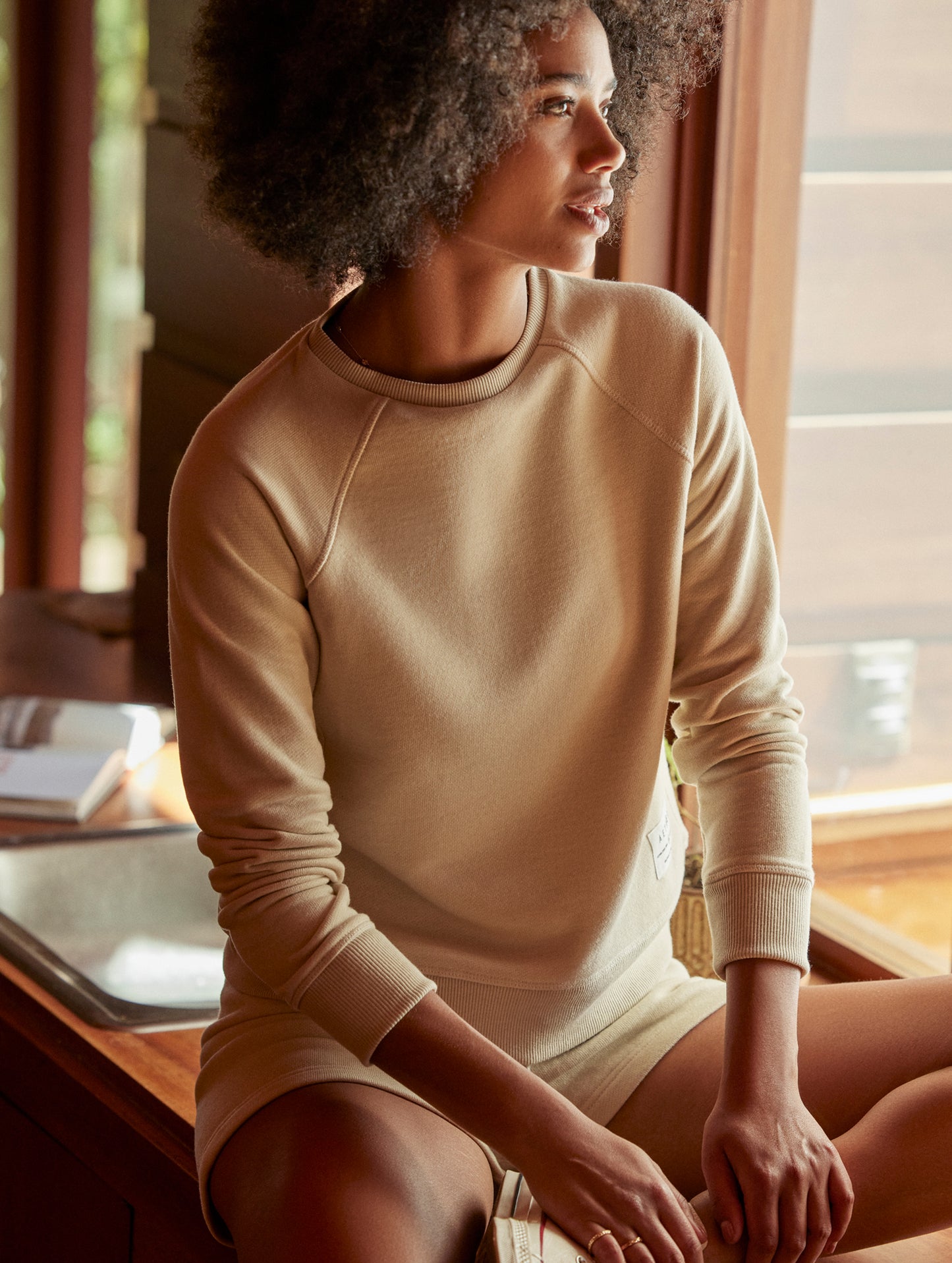 Front angled indoor lifestyle image of woman sitting on desk wearing Foundation Raglan Sweatshirt in Bone white from AETHER Apparel.